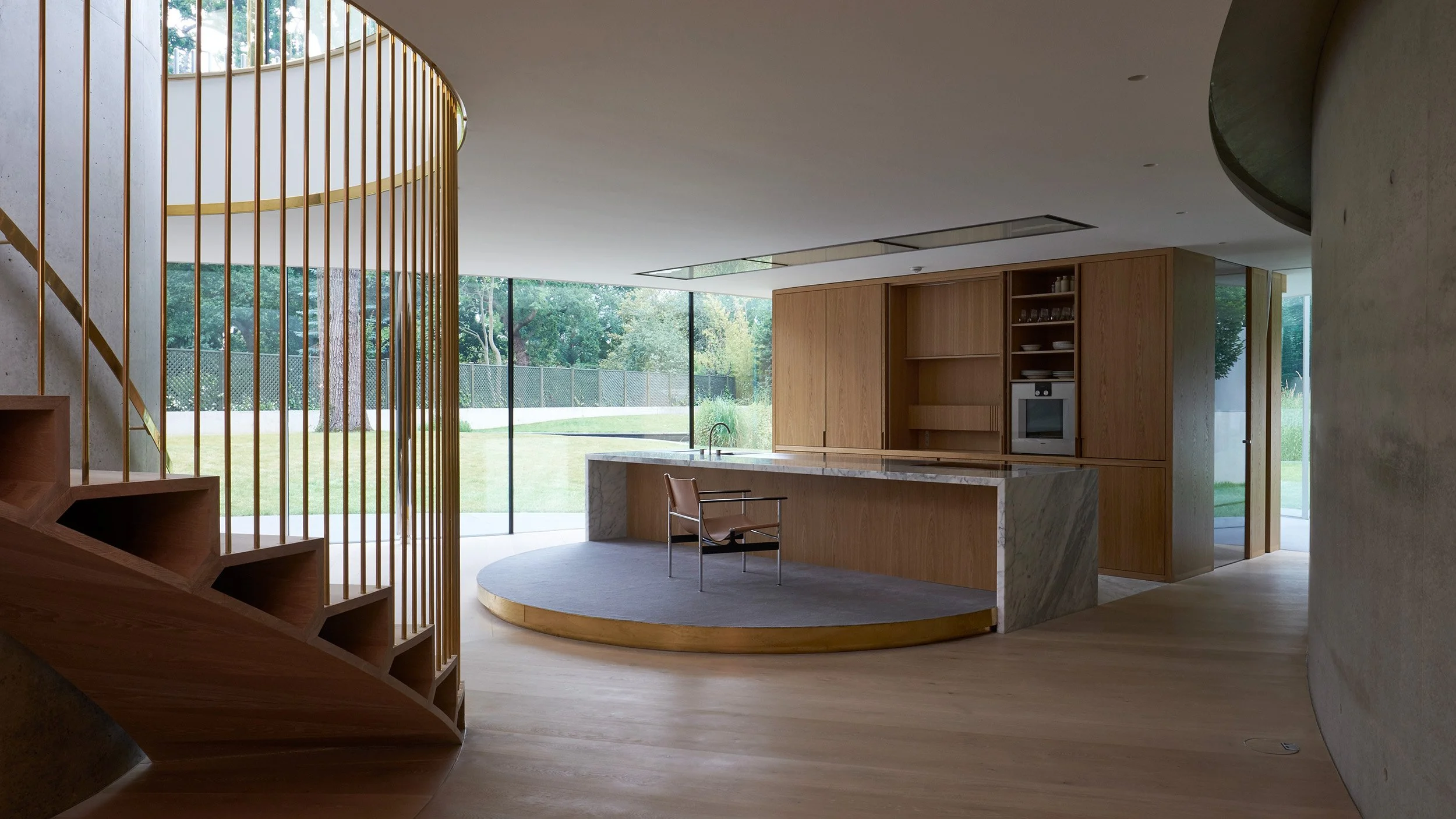 Modern kitchen interior with a curved wooden staircase, marble island, and large glass windows overlooking a garden.
