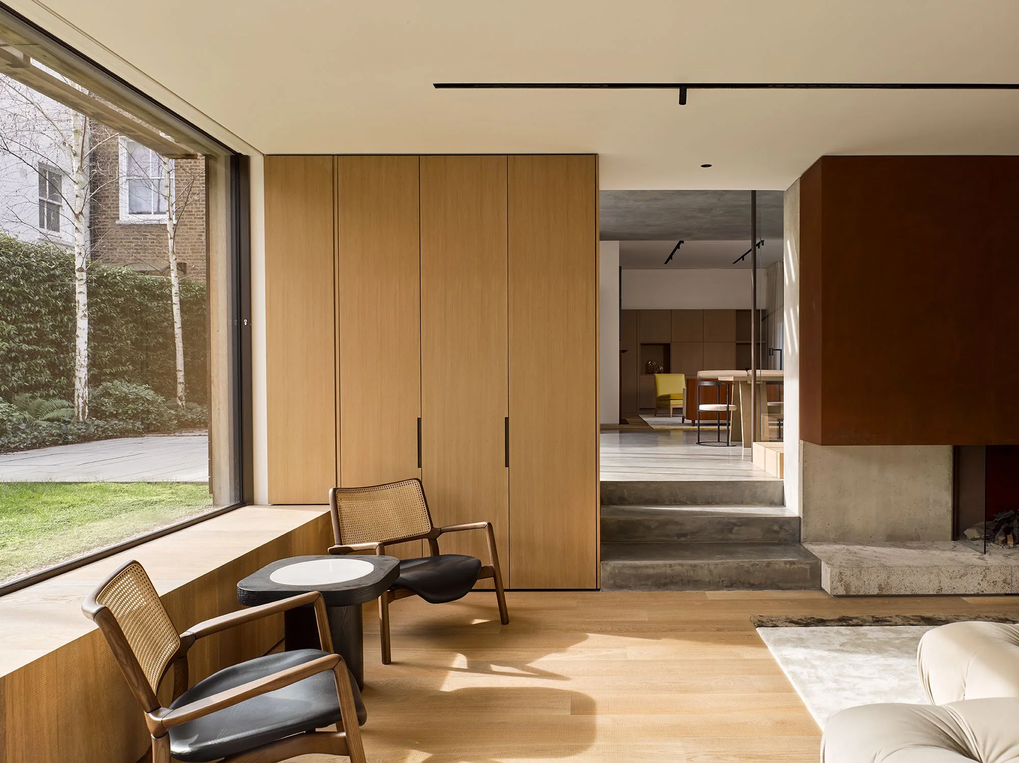 Modern living room interior with large window, two mid-century chairs, round side table, wooden floors, and a view of a garden outside. Adjacent room visible with dining area and neutral color palette.
