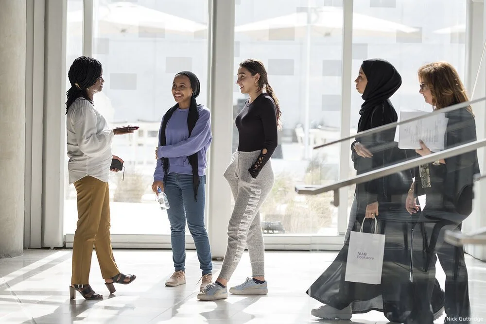 Northwestern University In Qatar. Five women in casual and business attire standing and talking inside a modern building with large windows.