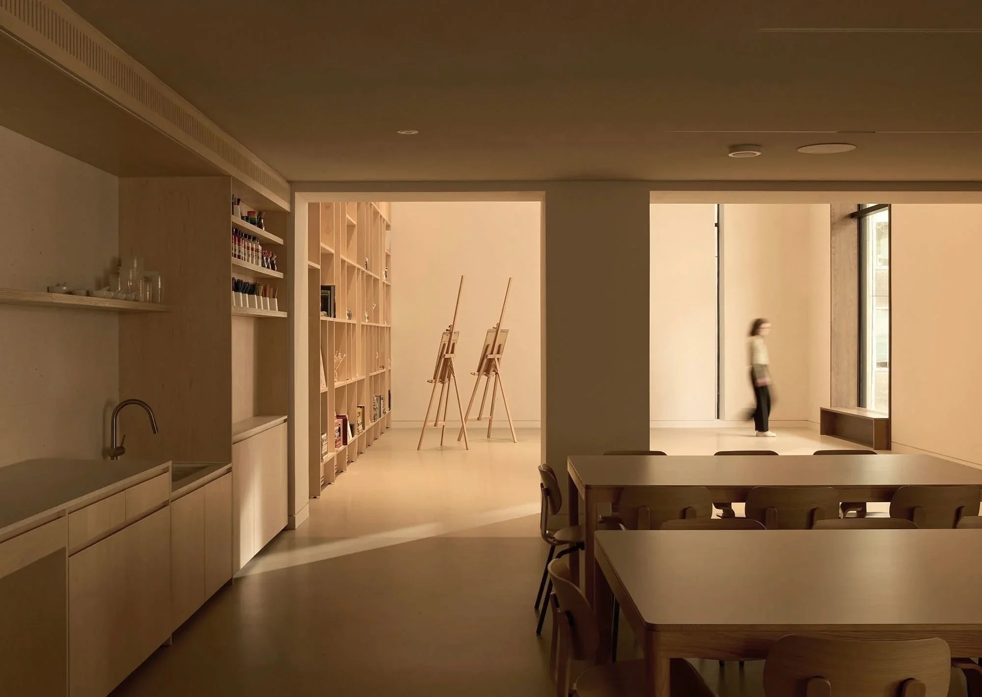 Interior of a modern art studio with tables, chairs, shelves holding art supplies, and a person walking by. Wooden easels are placed near shelves, which contain various art materials on display.
