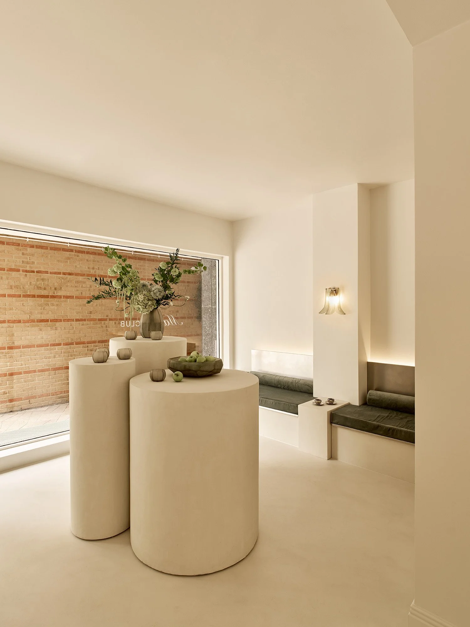 Onyx Matcha Club, London's new destination for matcha and modern rituals. Minimalist interior with white cylindrical tables, a vase with flowers on top, a brick wall with large window, and built-in seating with cushions, illuminated by wall sconces.