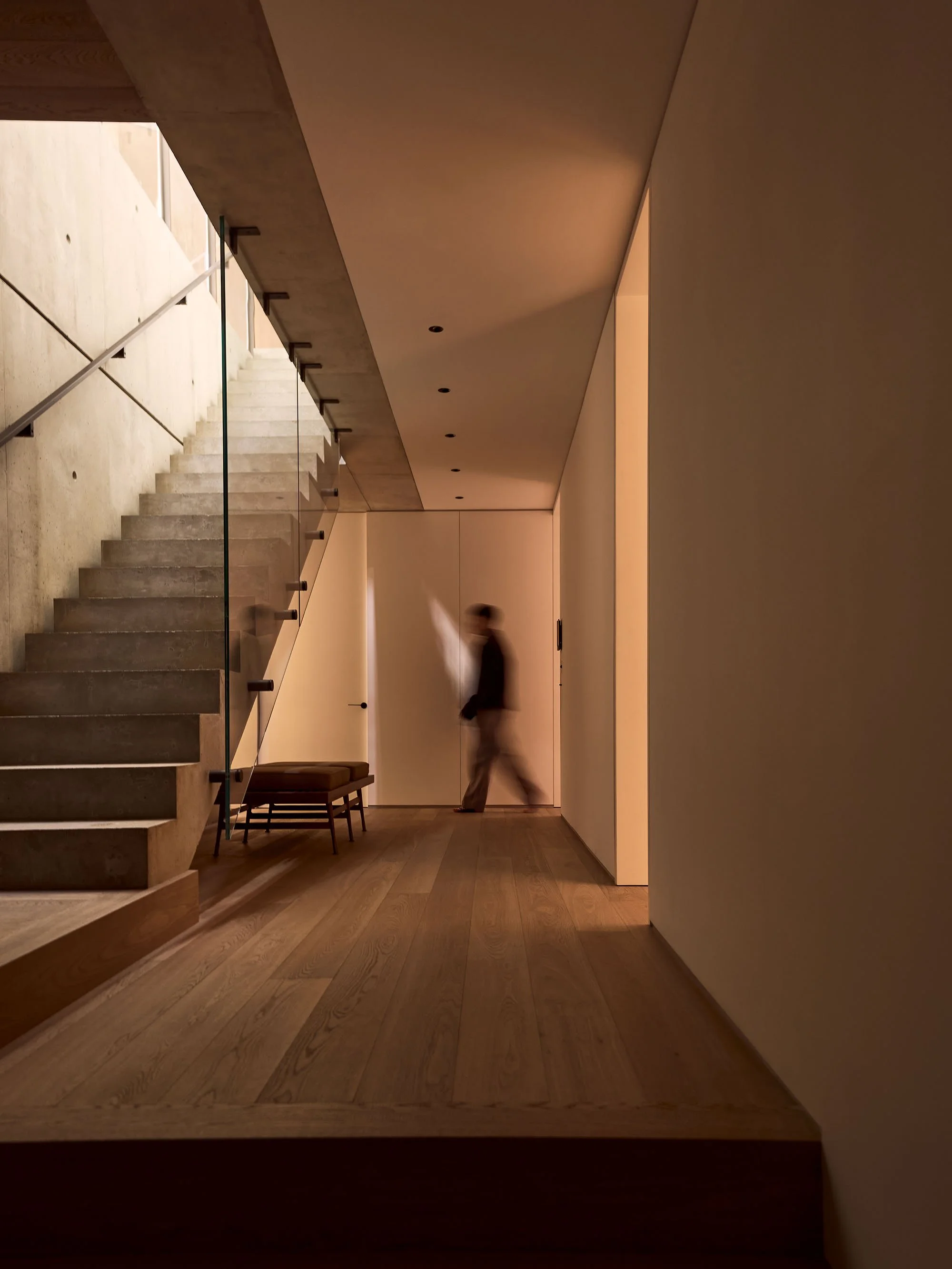 Modern interior with wooden flooring, concrete staircase, glass railing, and blurred person walking.