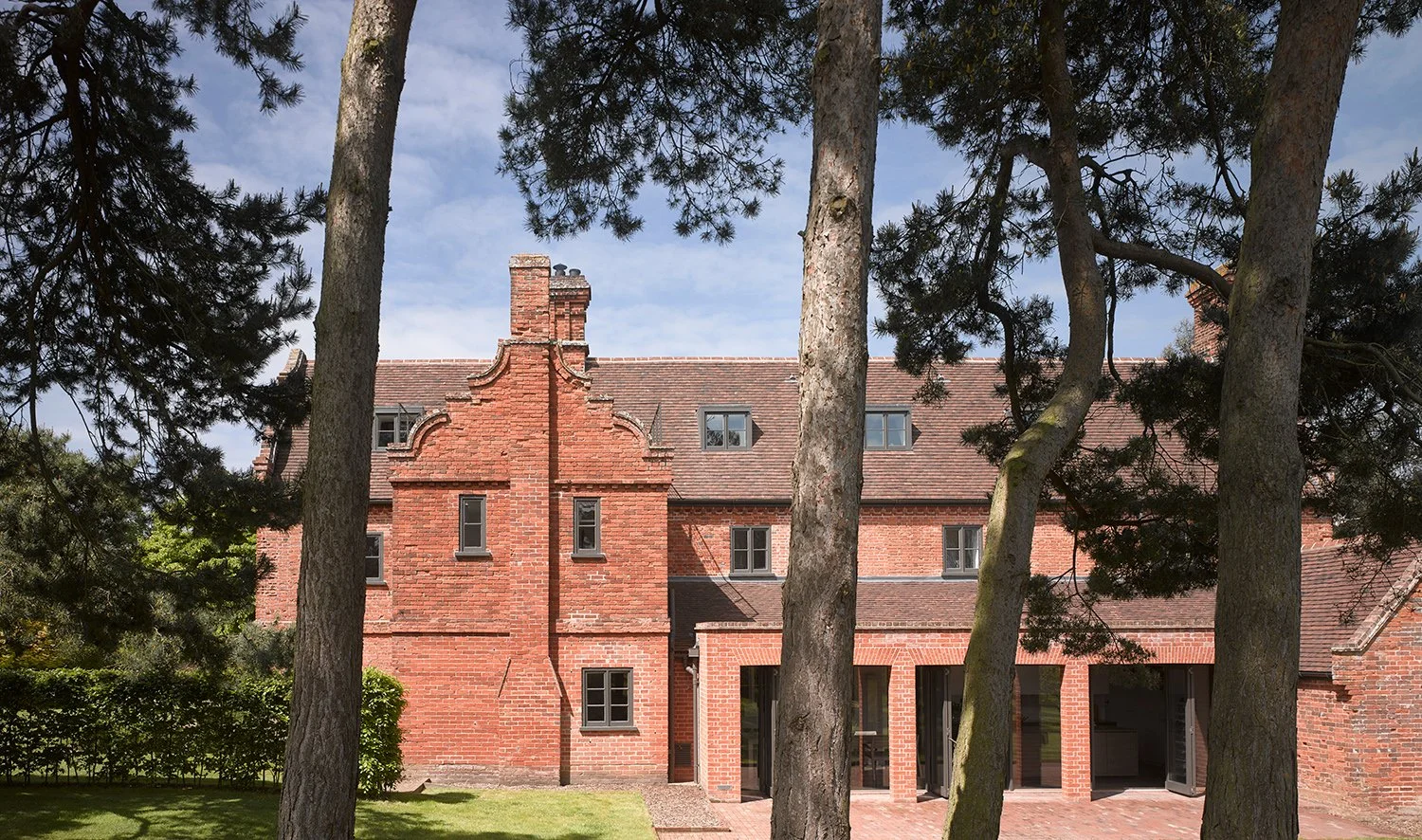 A red brick house partially obscured by tall trees with a brick patio in the backyard, under a partly cloudy sky.