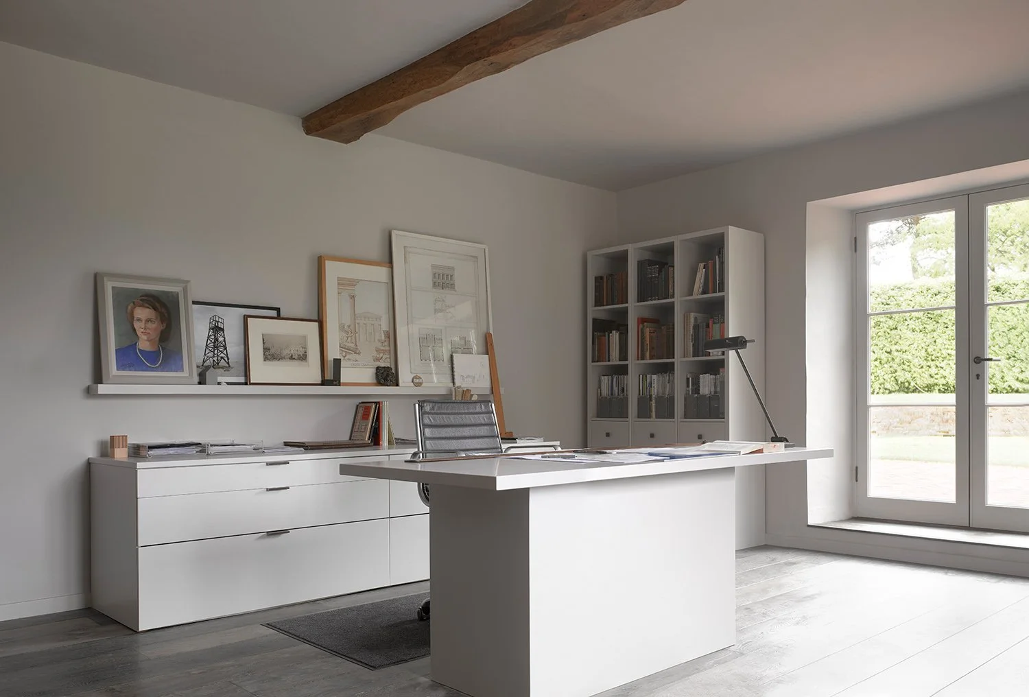 Modern home office with white furniture, bookshelf, framed artwork, and a glass door leading outside, illuminated by natural light.