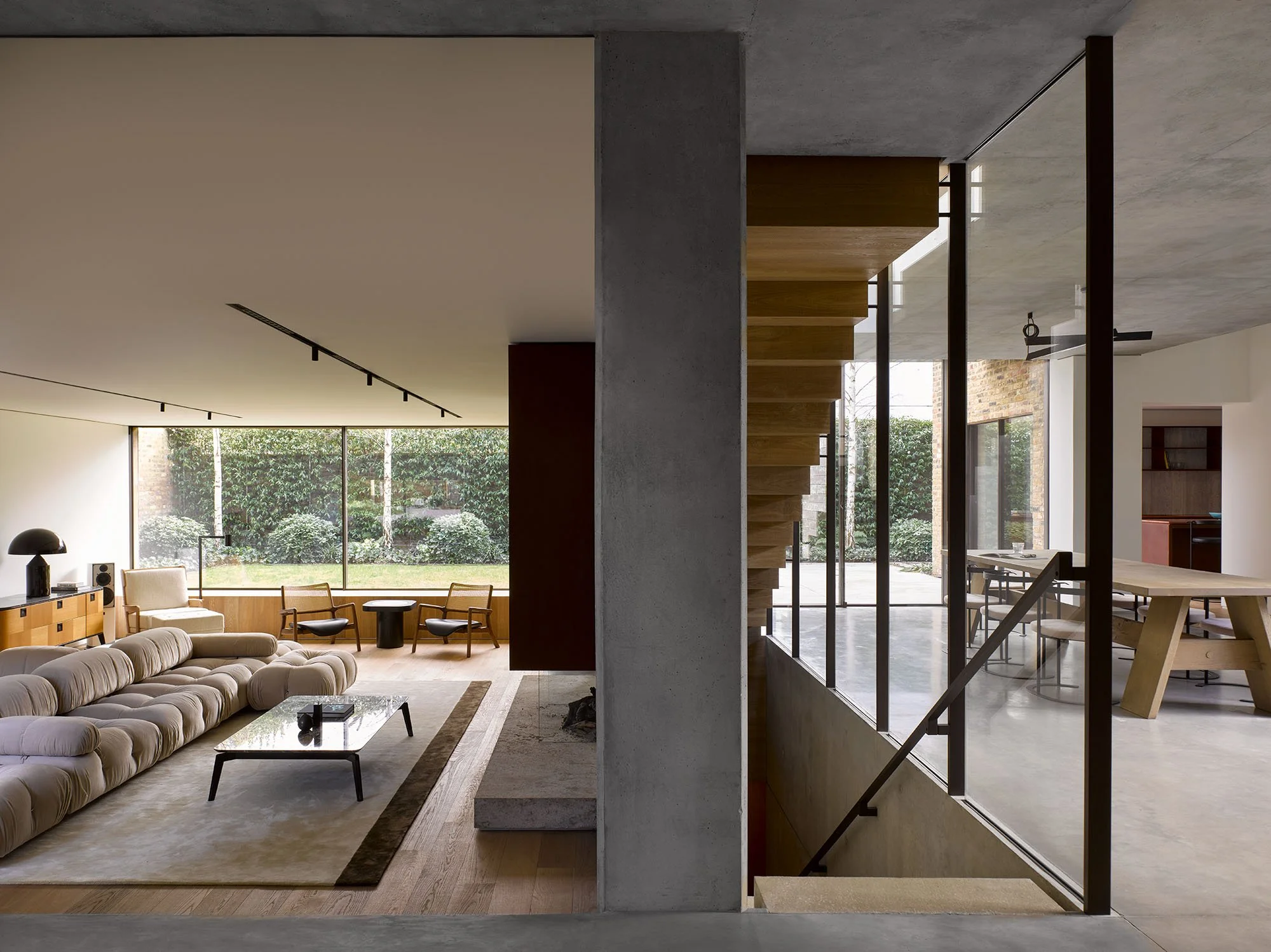 Modern interior showcasing a living area with a plush sofa, glass coffee table, and large window overlooking greenery. Adjacent open-space features a wooden staircase and dining area with a long table. The design emphasizes natural light and minimali