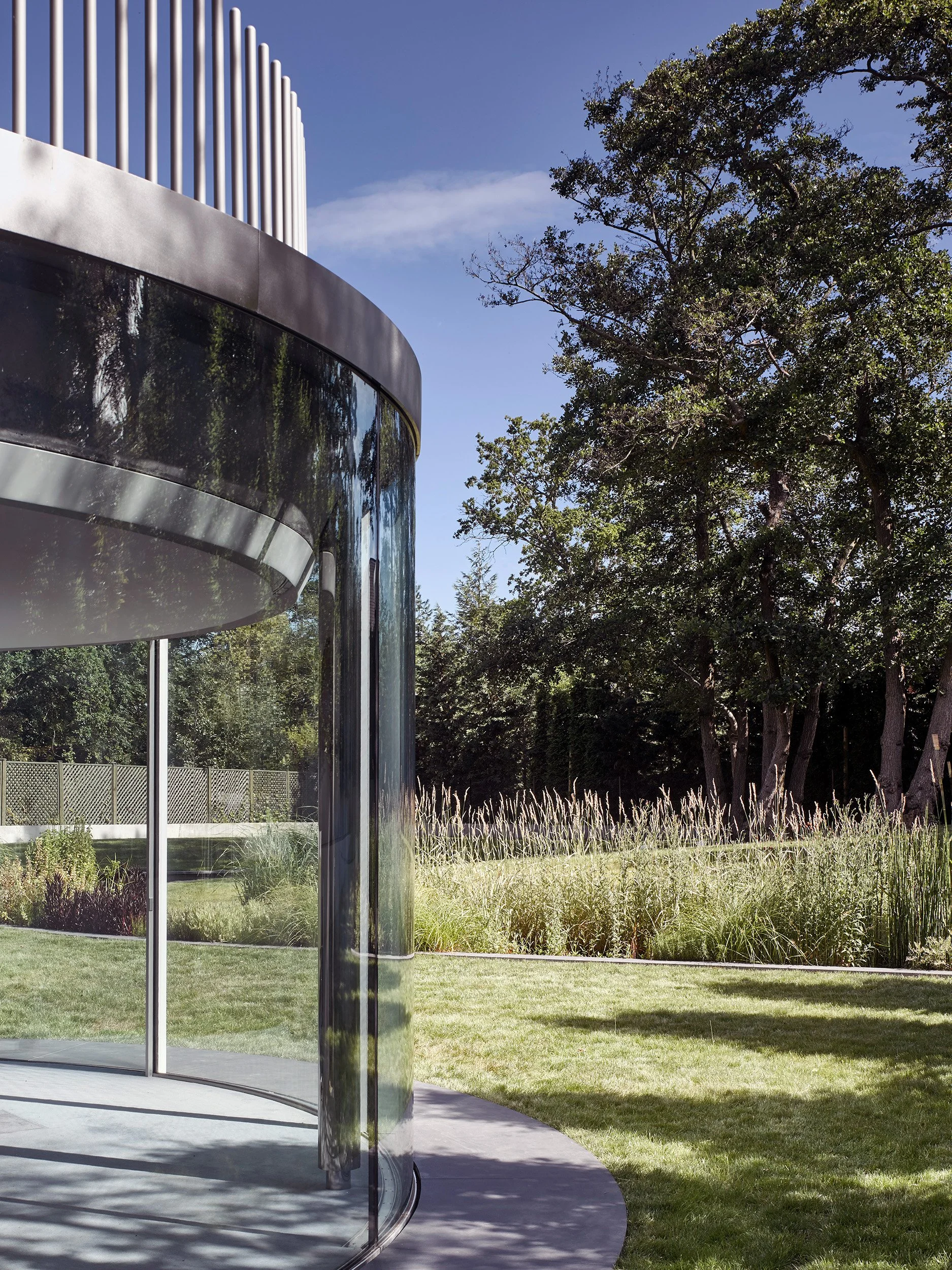 Modern circular glass building beside grassy area and tall trees under a clear blue sky.
