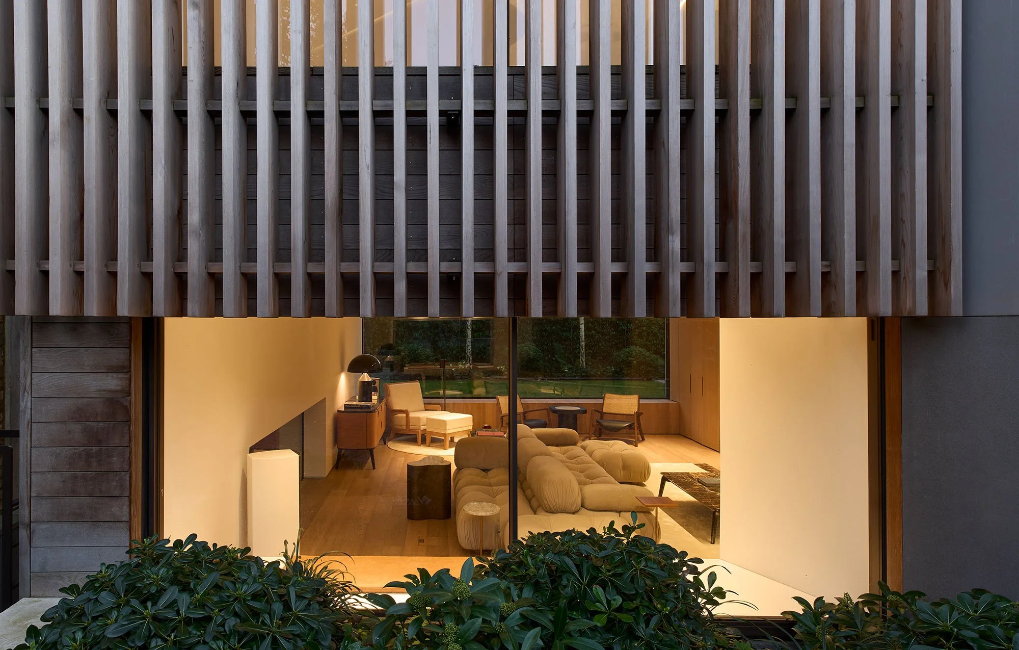 Modern living room with large windows and wooden slat exterior, featuring a cozy sofa, chairs, and a coffee table, view from outside with greenery.