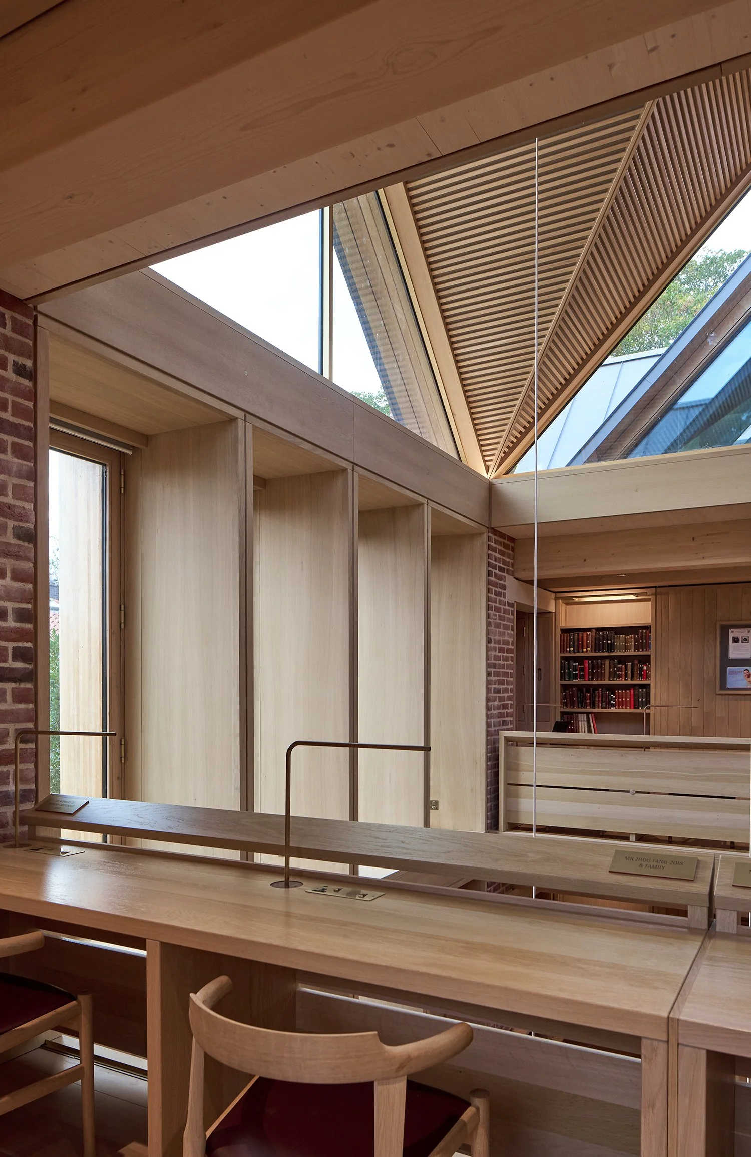 Interior of a modern library with wooden desks and chairs, glass windows, and a collection of books on shelves.