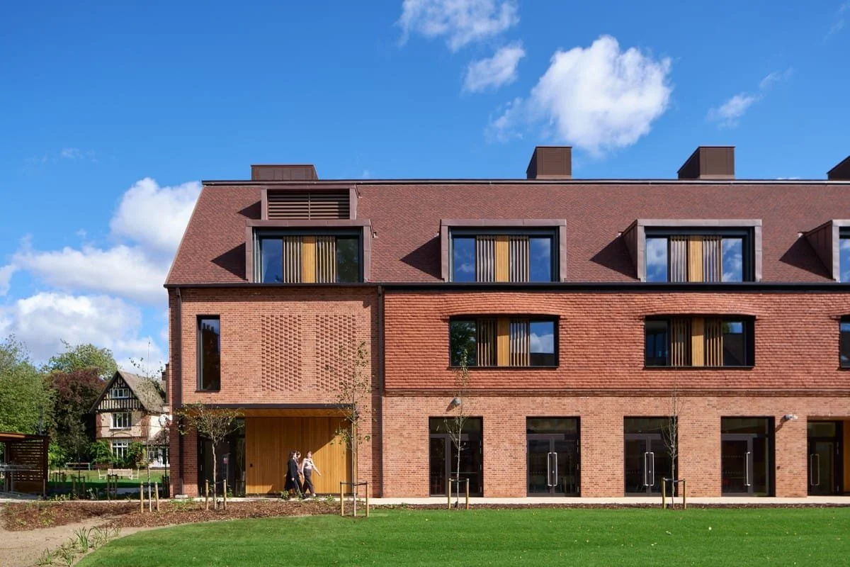 Modern brick apartment building with large windows, set against a blue sky with scattered clouds, with a green lawn in front and people walking.