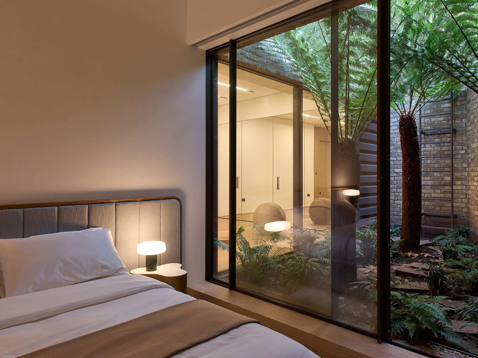 Modern bedroom with a large window view of an outdoor garden featuring lush ferns and trees. The room has a minimalist bed with white bedding and a small bedside table with a lamp.