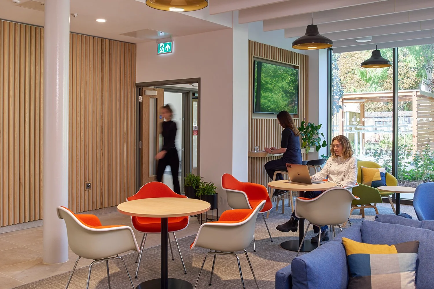 Interior of a modern lounge or café with colorful chairs, a woman working on a laptop, a person reading near a large window, and a staff member walking in the background.