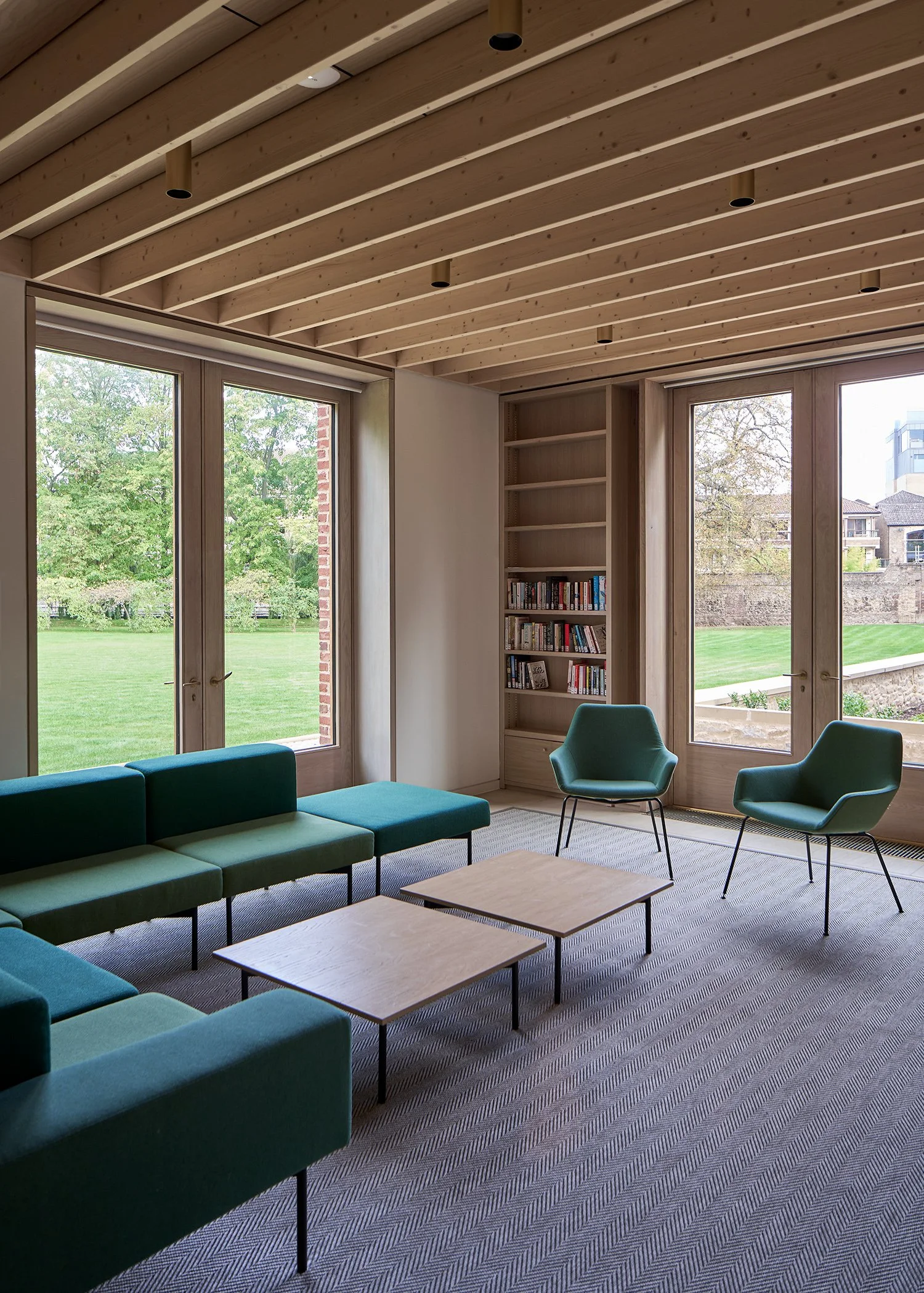 Modern living room with green sectional sofa, two green armchairs, wooden coffee tables, and built-in wooden shelving with books. Large windows overlook a green lawn.
