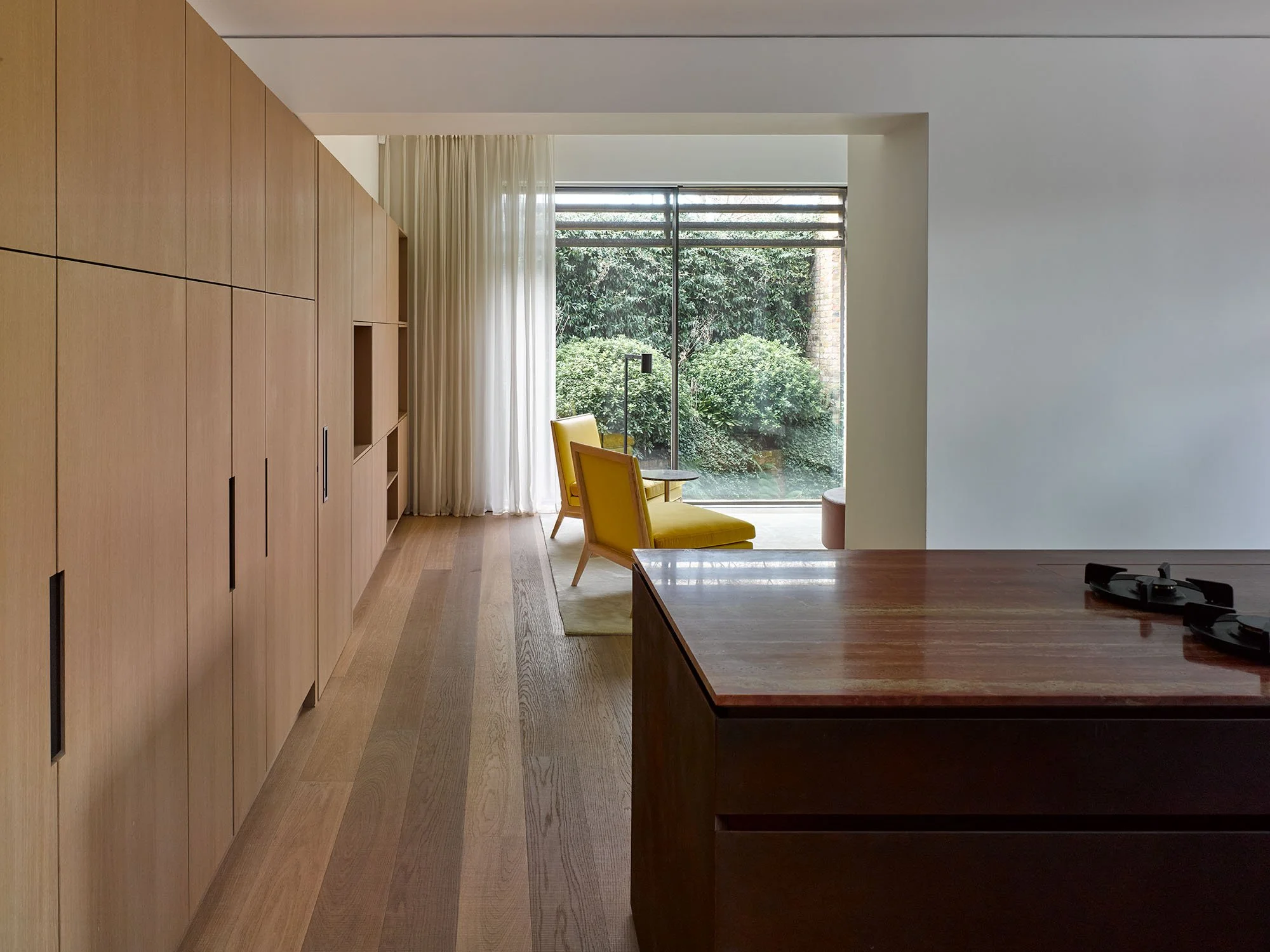 Modern interior with light wood cabinets, a wooden floor, and yellow armchairs near a large glass window overlooking greenery.