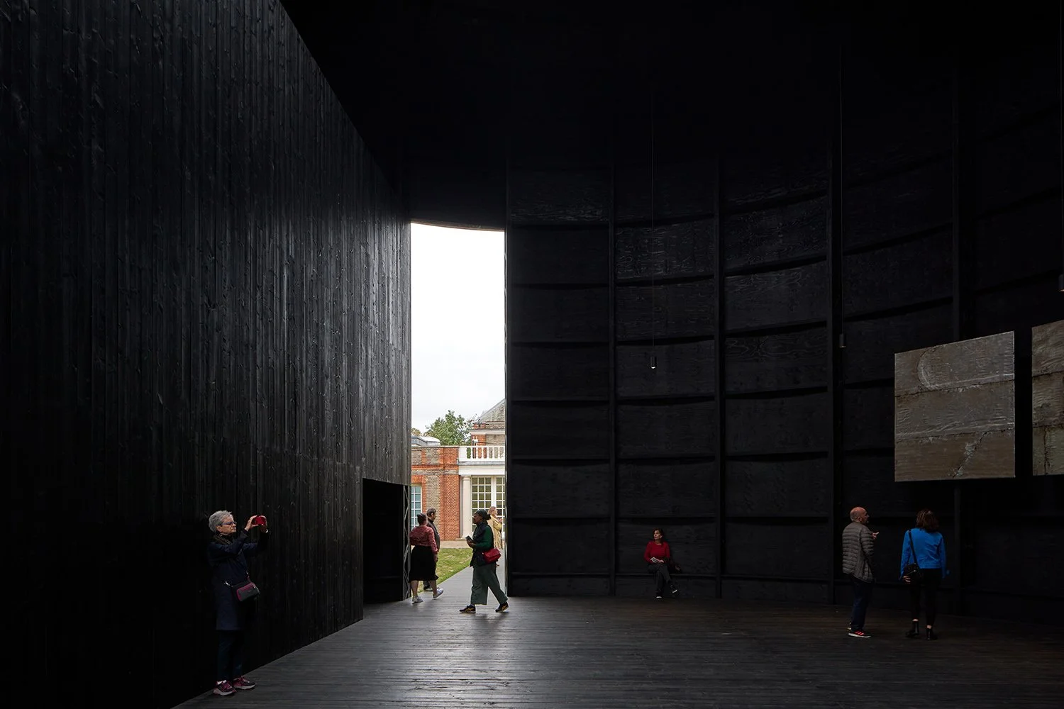 Interior of a dark, minimalist architectural space with textured black walls and wooden flooring. Several people are inside, some taking pictures and others observing the surroundings. A bright opening shows the outside with a glimpse of a brick buil