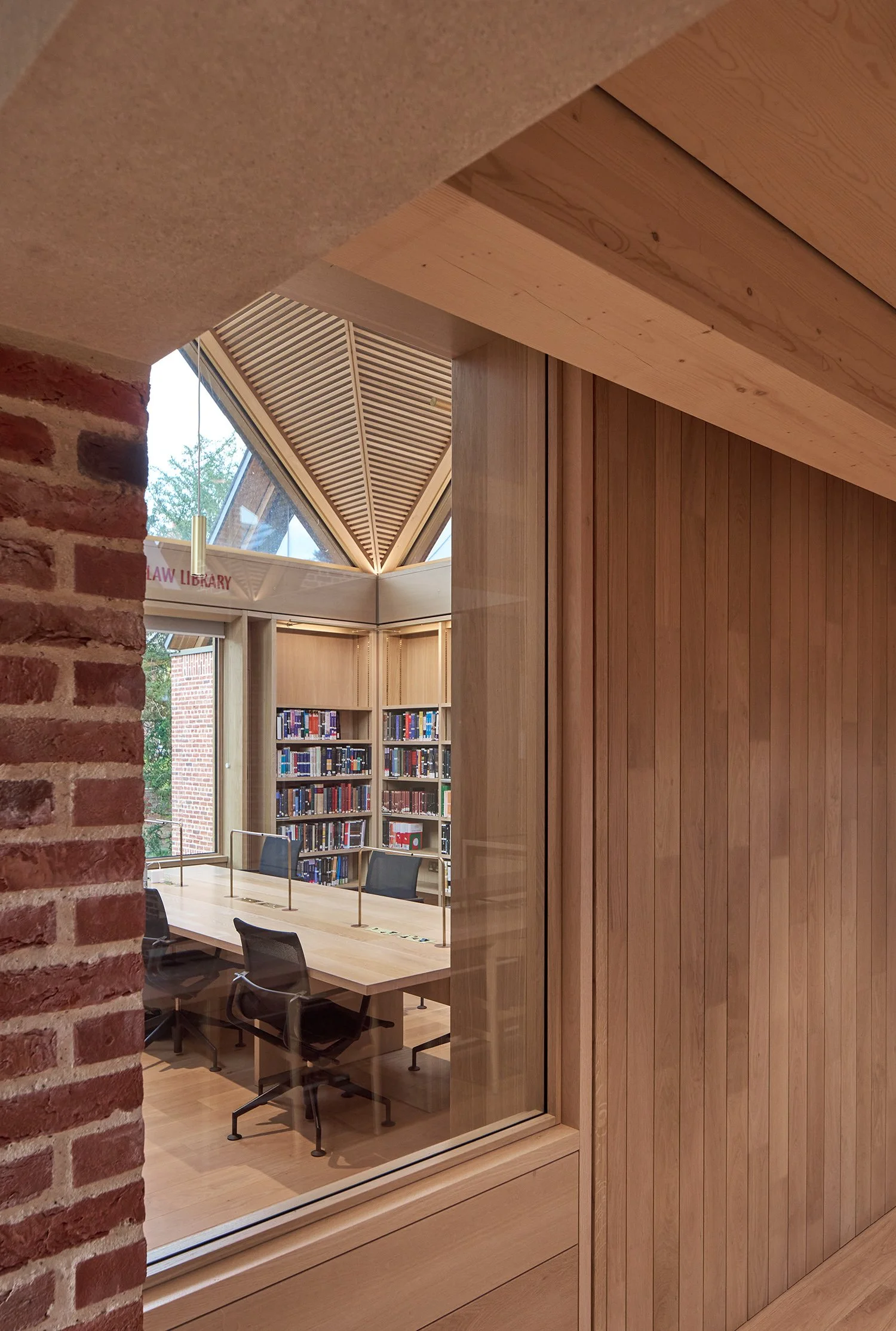Interior of a modern library with wooden paneling, bookshelves filled with books, and a conference table with chairs. Large glass windows and a slanted roof ceiling provide natural light.