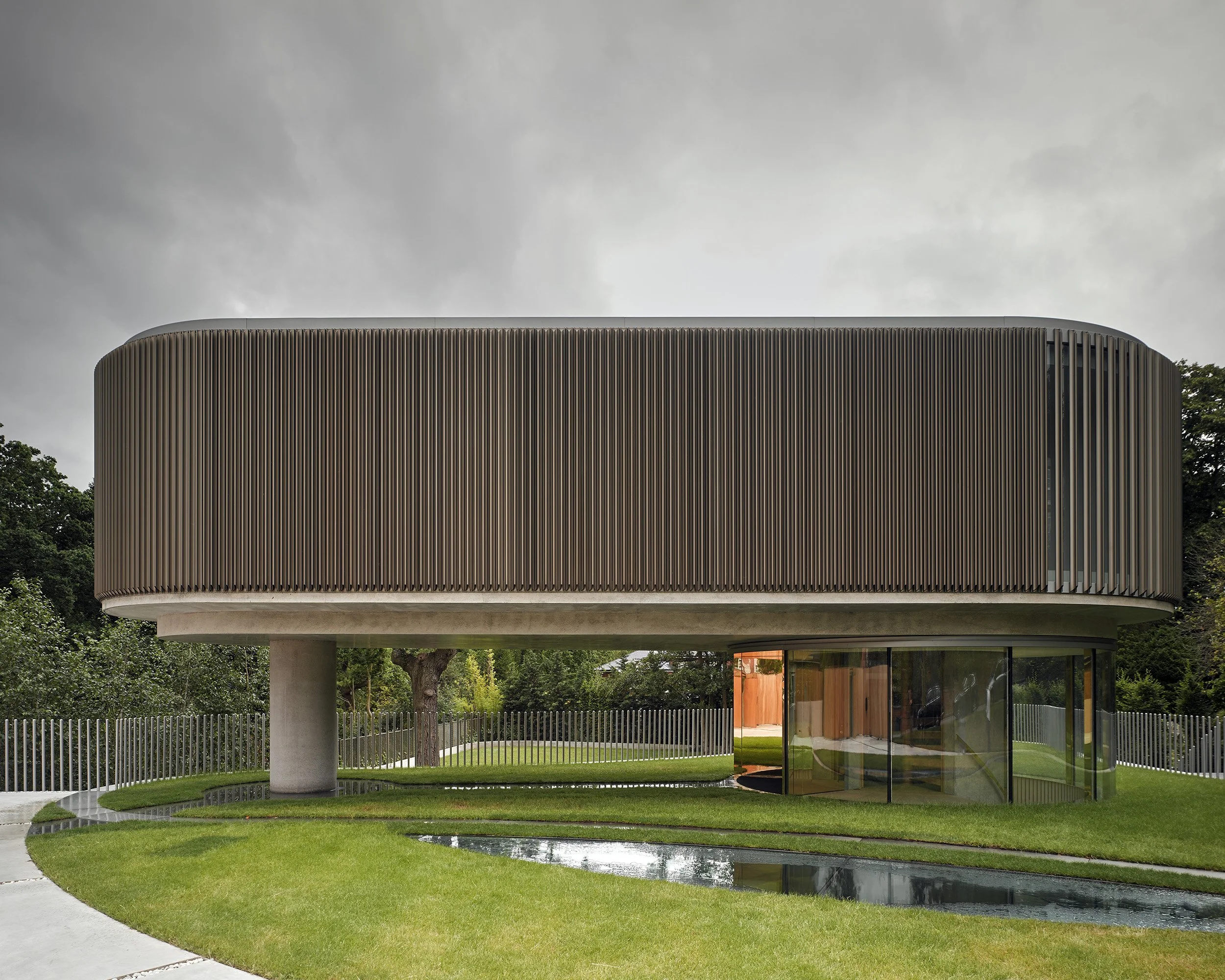Modern cylindrical building with vertical slats and glass walls, elevated above the ground on a central column, surrounded by a landscaped garden with a curved pathway and water feature.