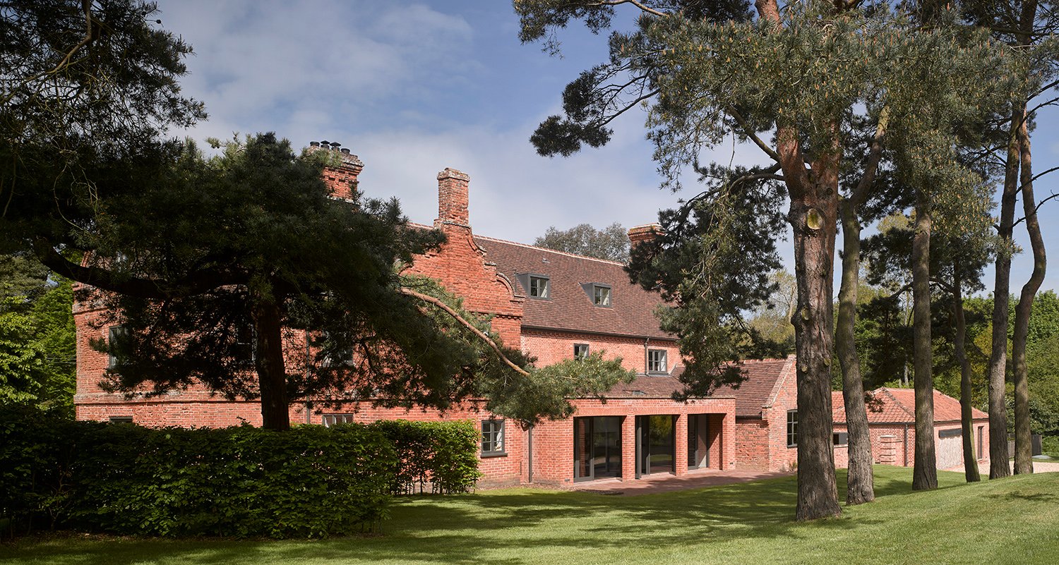 Red brick house with multiple chimneys, large windows, surrounded by trees and a grassy lawn.