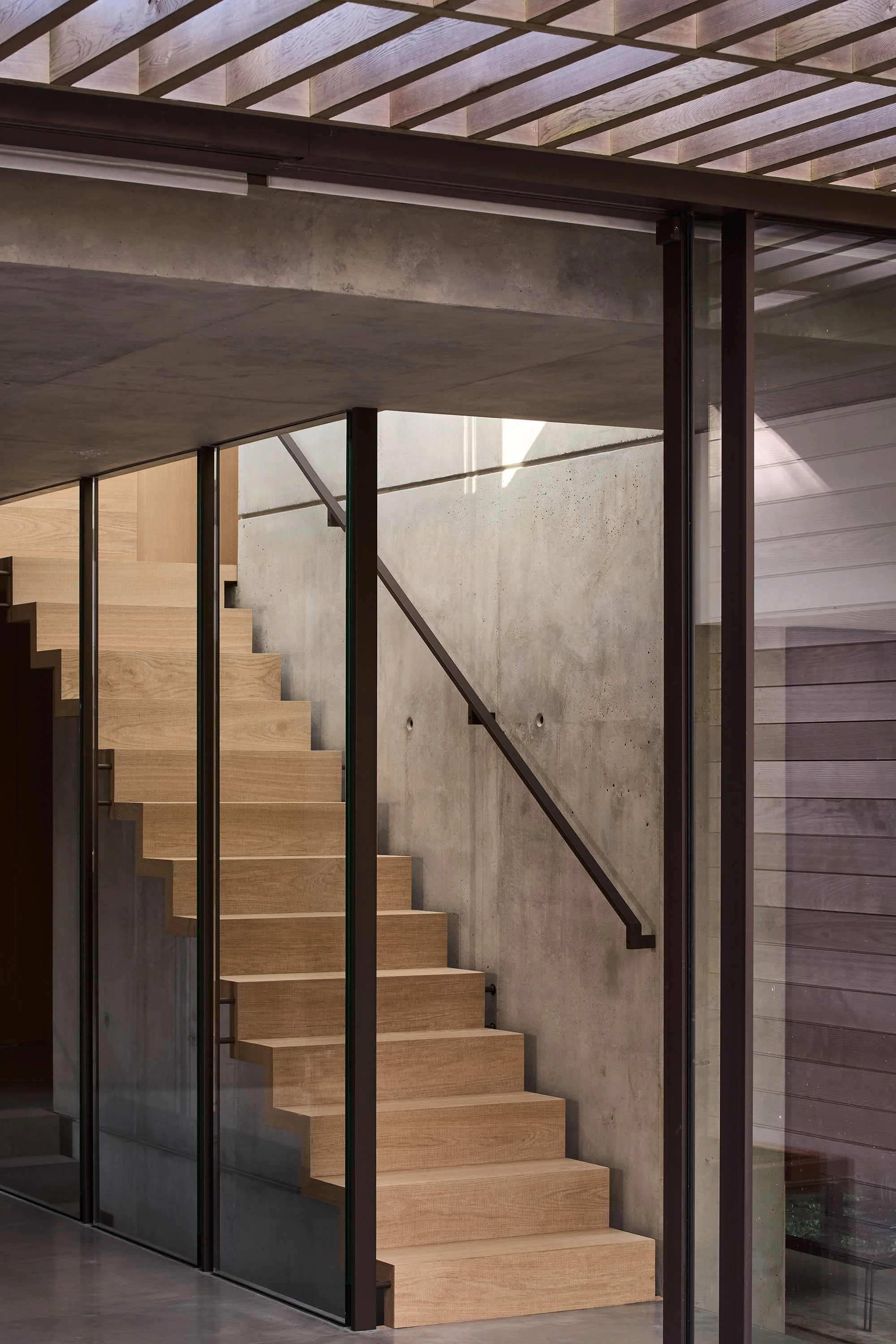Modern wooden staircase with glass walls and metal railings, concrete walls, and ceiling beams.