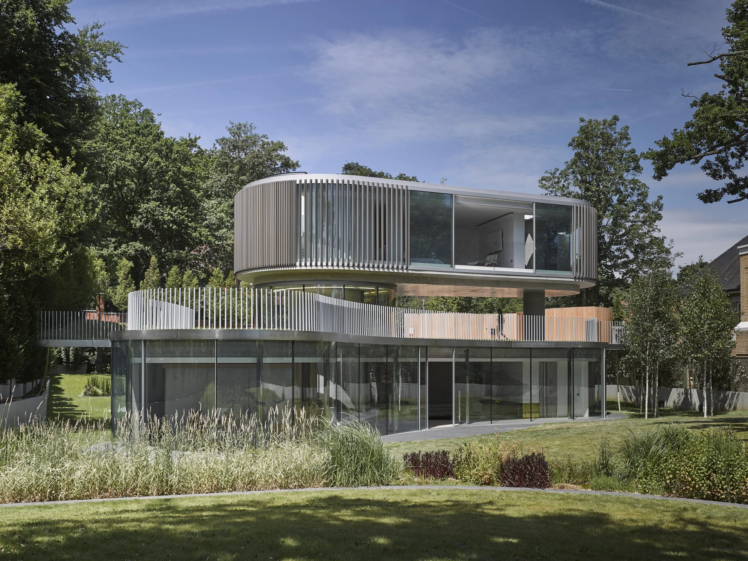 Modern house with unique, elevated circular structure, glass walls, surrounded by lush greenery and trees.