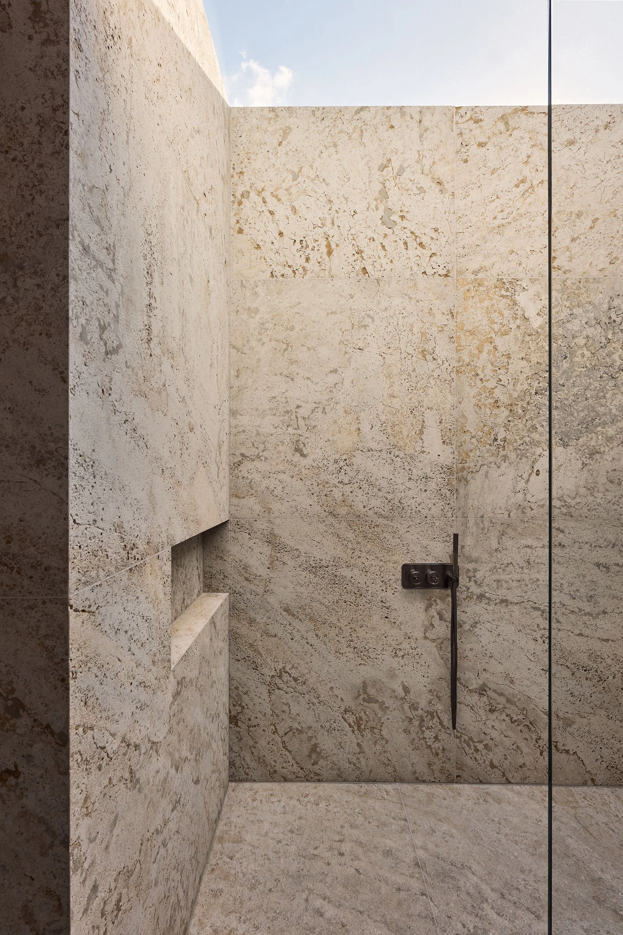 Minimalist stone shower with large tiles, built-in shelf, and metal fixtures under natural light.
