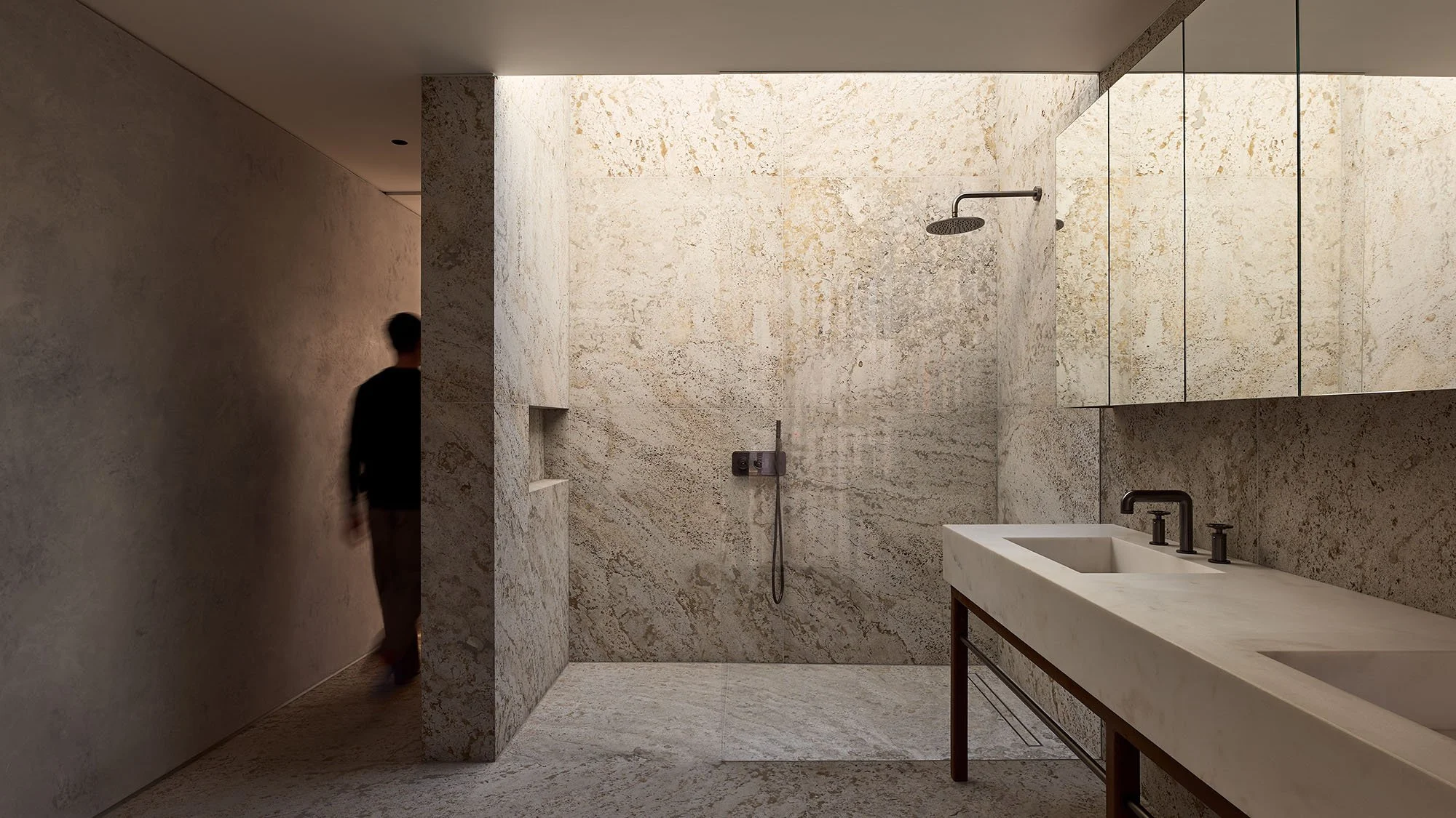 Modern bathroom with stone walls, double sink, and walk-in shower, featuring a person leaving the room.
