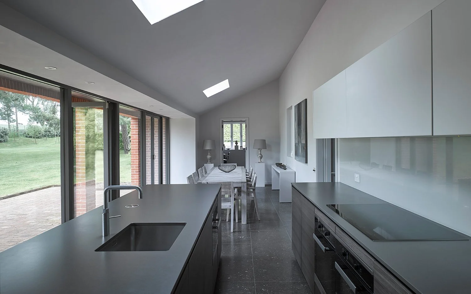 Modern kitchen with dark countertops, white cabinets, a large island with a sink, and a dining area with a table and chairs, adjacent to large glass sliding doors overlooking a green yard.
