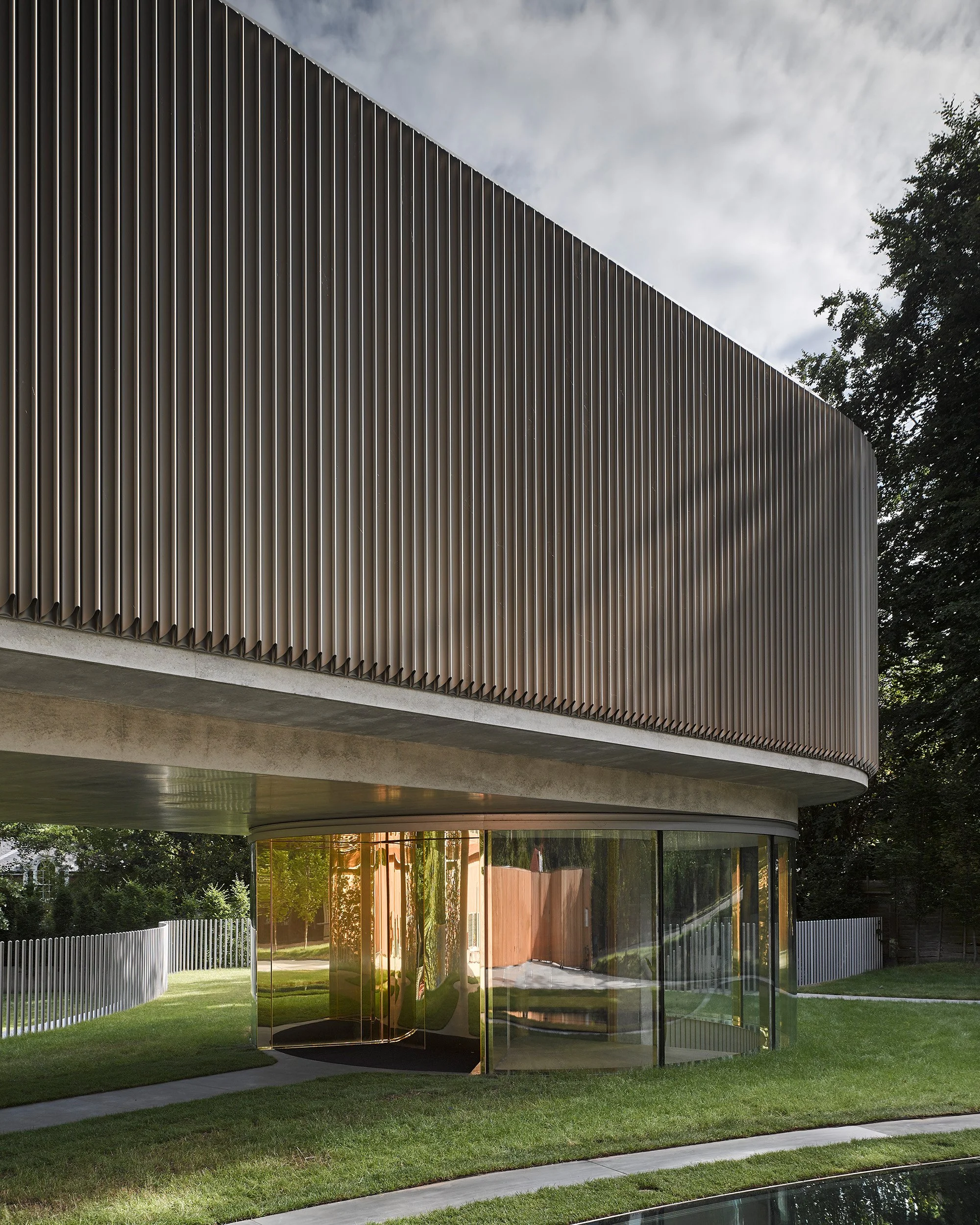 Modern architectural structure with vertical metal slats and glass walls, set in a landscaped area with grass and a curved pathway.