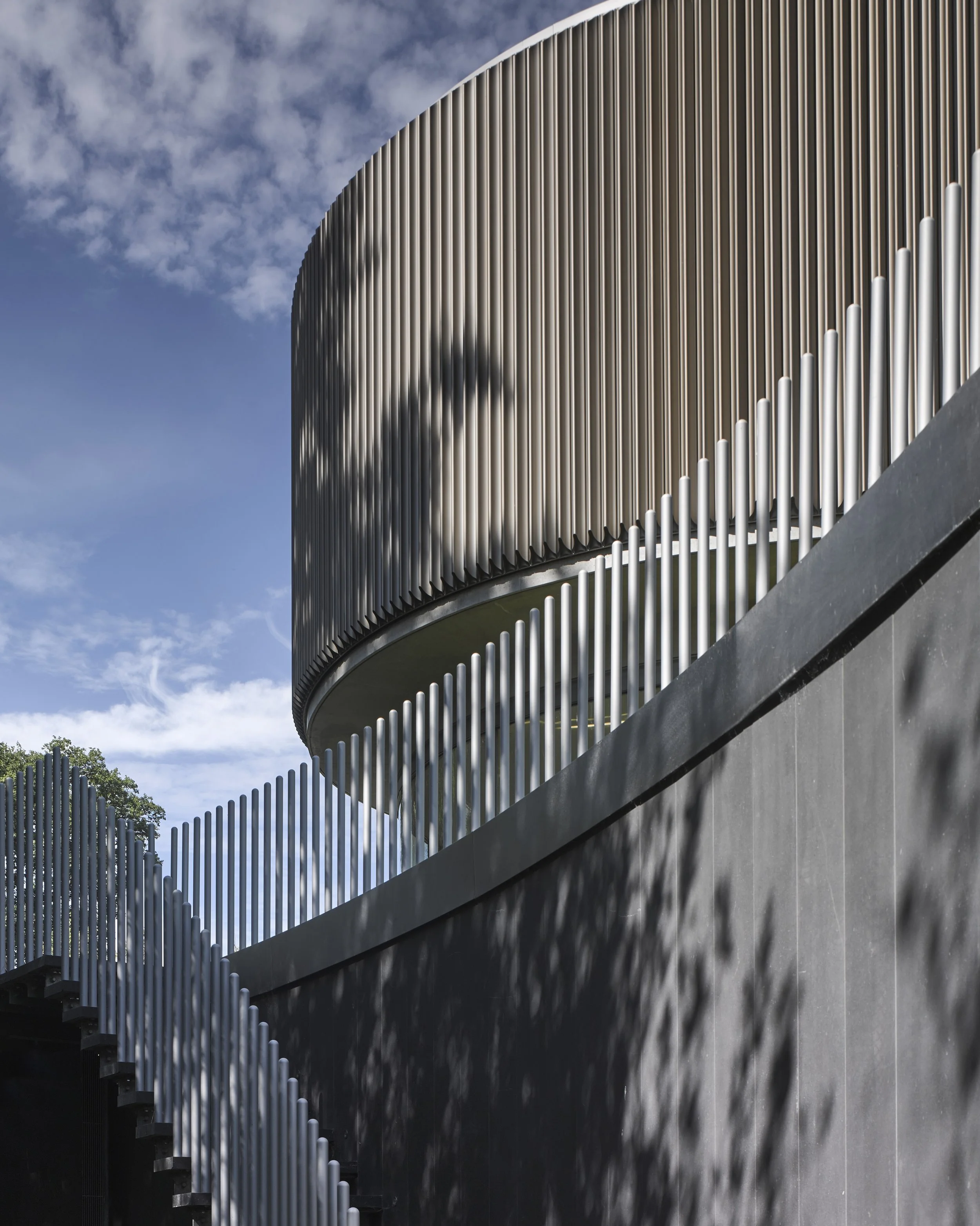 Modern architectural structure with vertical metal slats, curved walls, and shadows, viewed from below against a blue sky with scattered clouds.
