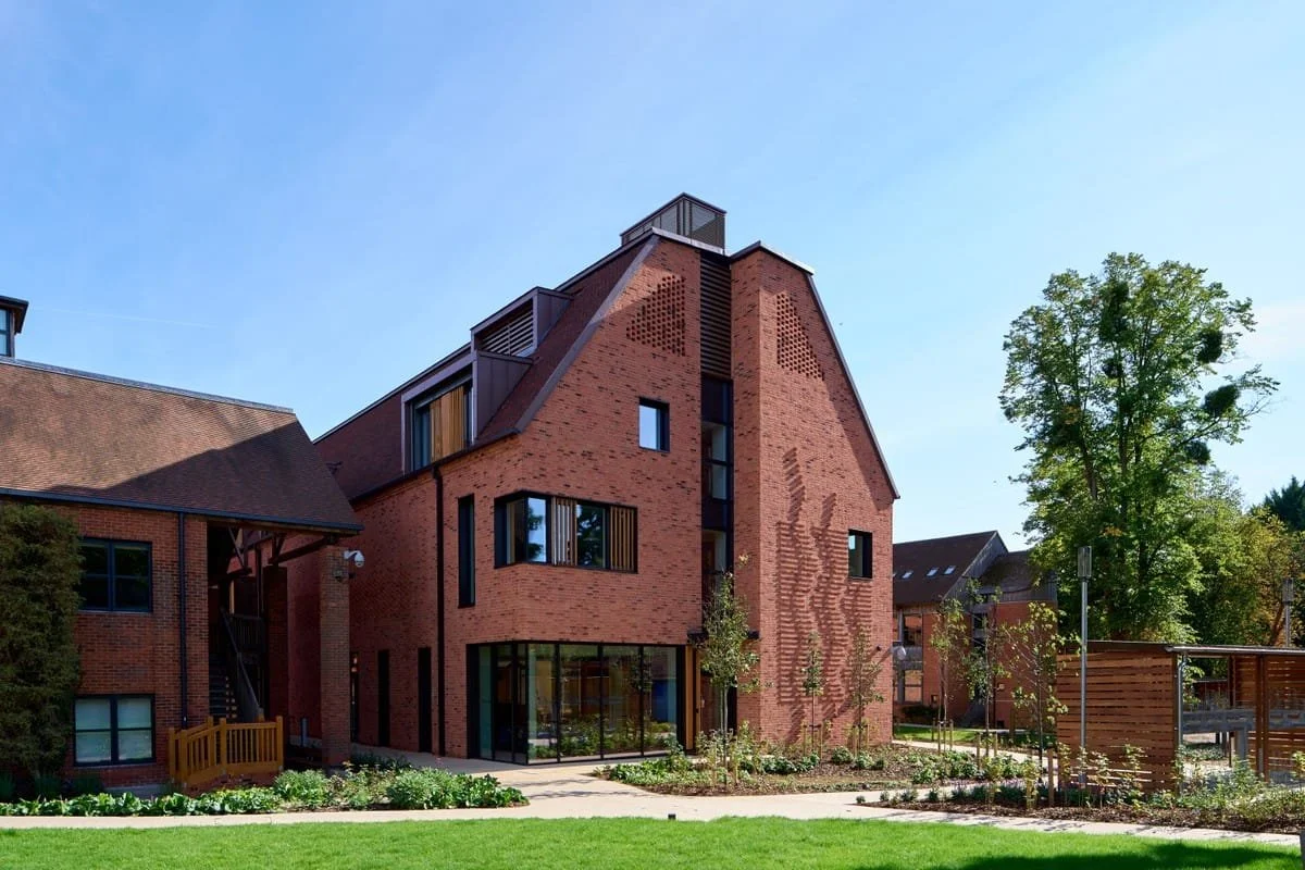 A modern red brick building with large windows and multiple levels, surrounded by a landscaped garden with grass, trees, and pathways.