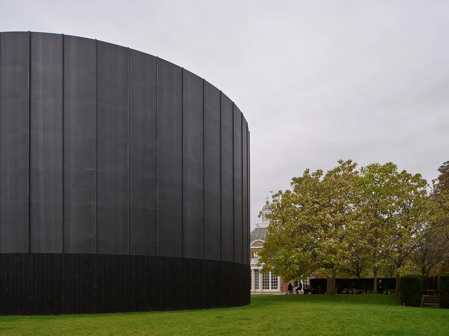 Large cylindrical building with black facade next to trees and a grassy area, featuring a traditional brick building in the background.