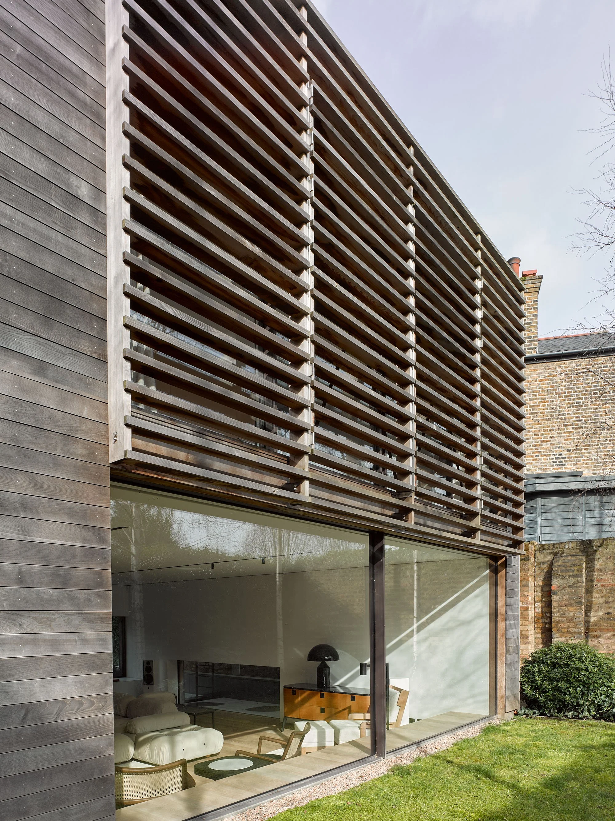 Modern house exterior with wooden slatted facade and large glass windows showcasing interior furniture like a sofa and lamp, adjacent to a garden lawn.
