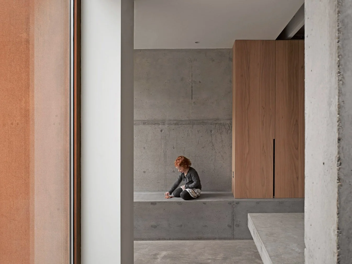 A woman with red hair sitting on a concrete ledge in a modern, minimalist interior with concrete walls, wooden panel, and large windows.