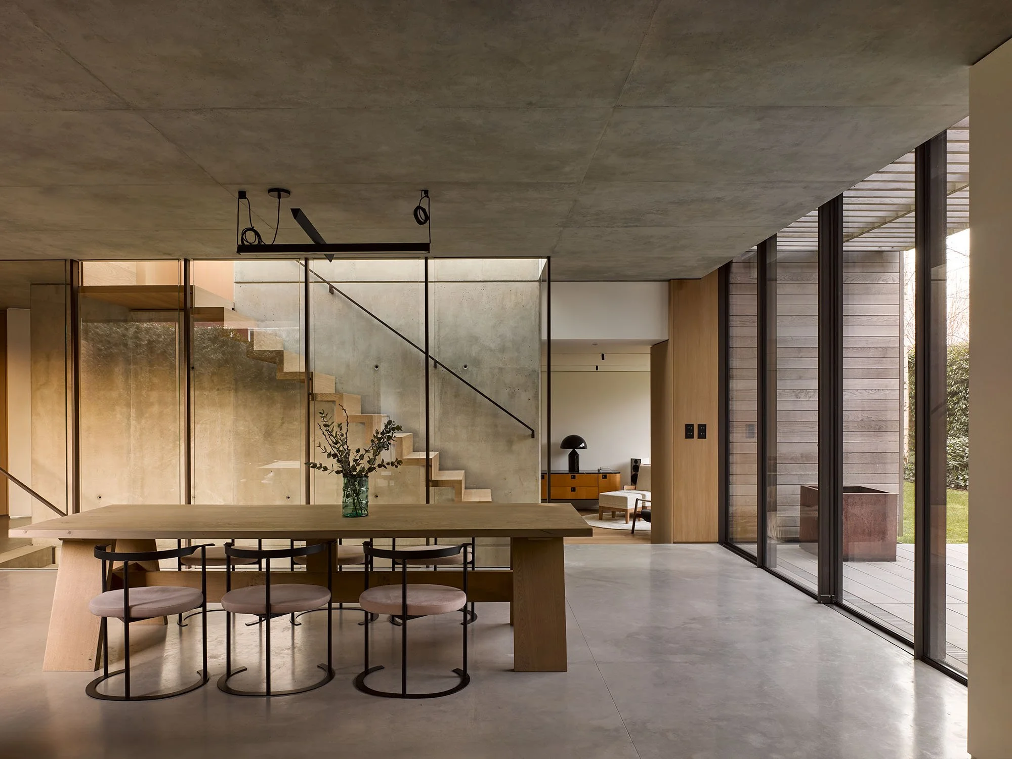 Modern interior with concrete walls, glass staircase, wooden dining table, round chairs, floor-to-ceiling windows, and a minimalist decor.