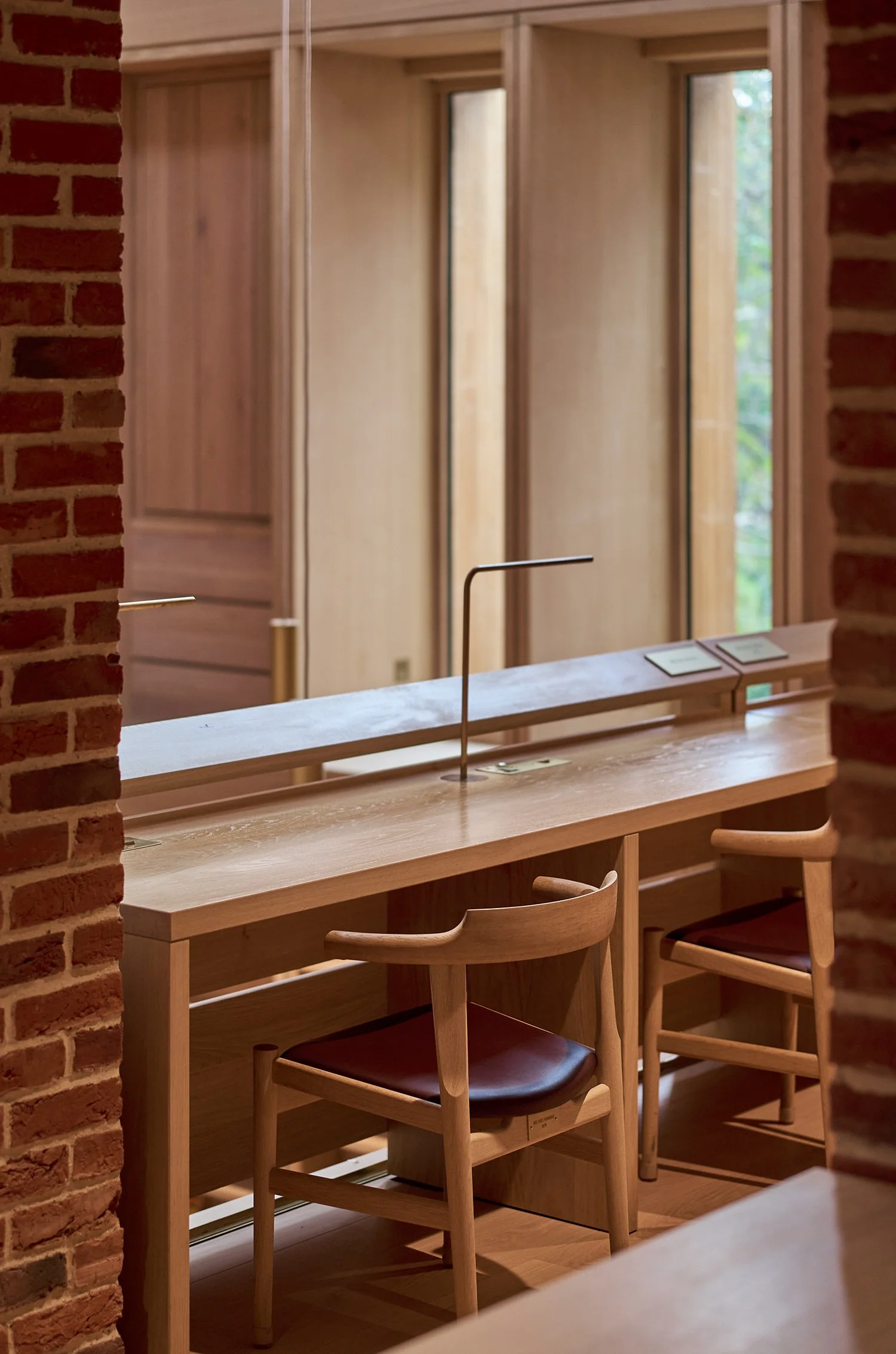 Modern library study area with wooden desks and chairs, minimalist lamps, and large windows framed by brick walls.