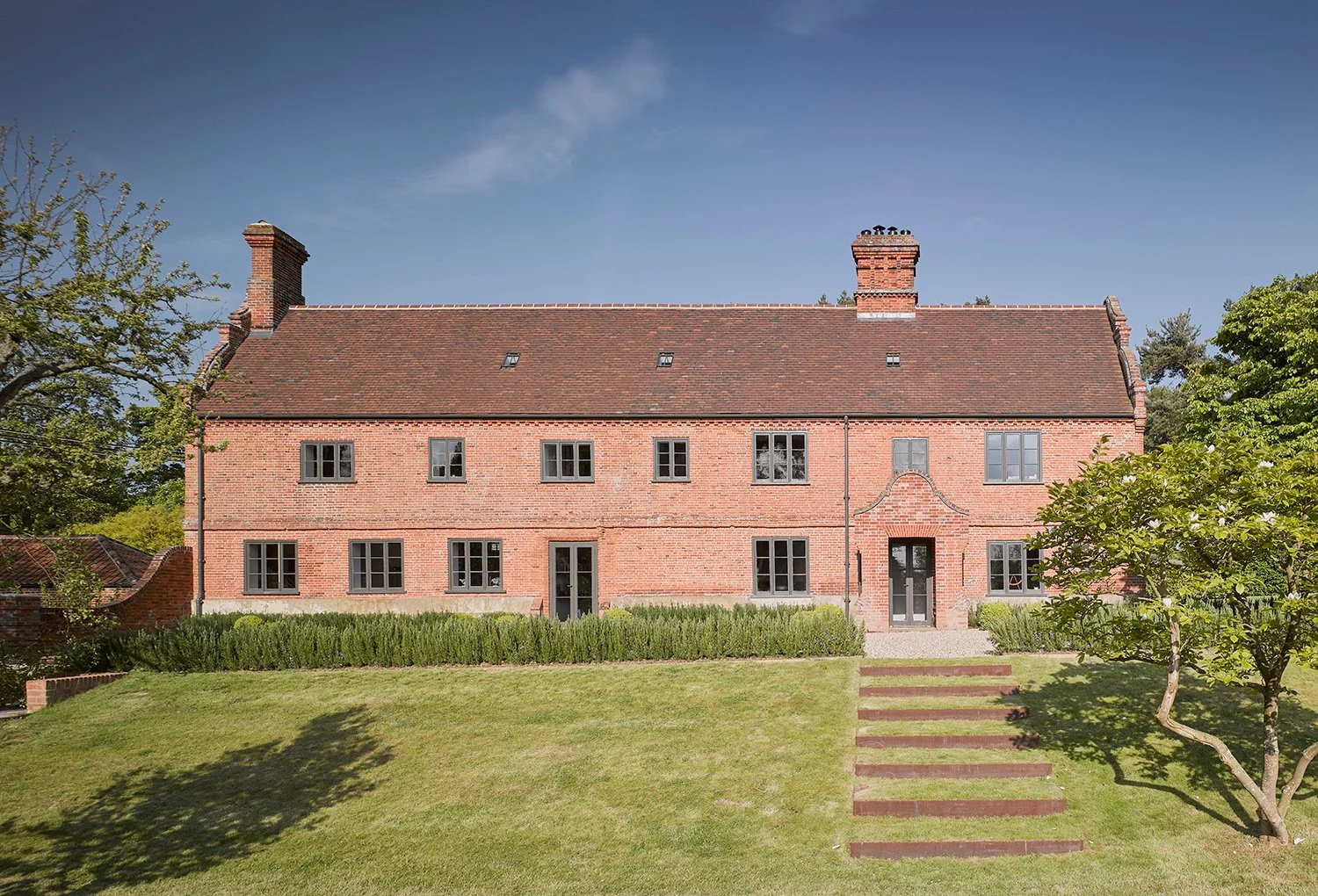 A large red brick house with a sloped roof, multiple windows, three chimneys, and a garden with a pathway and trees in front.
