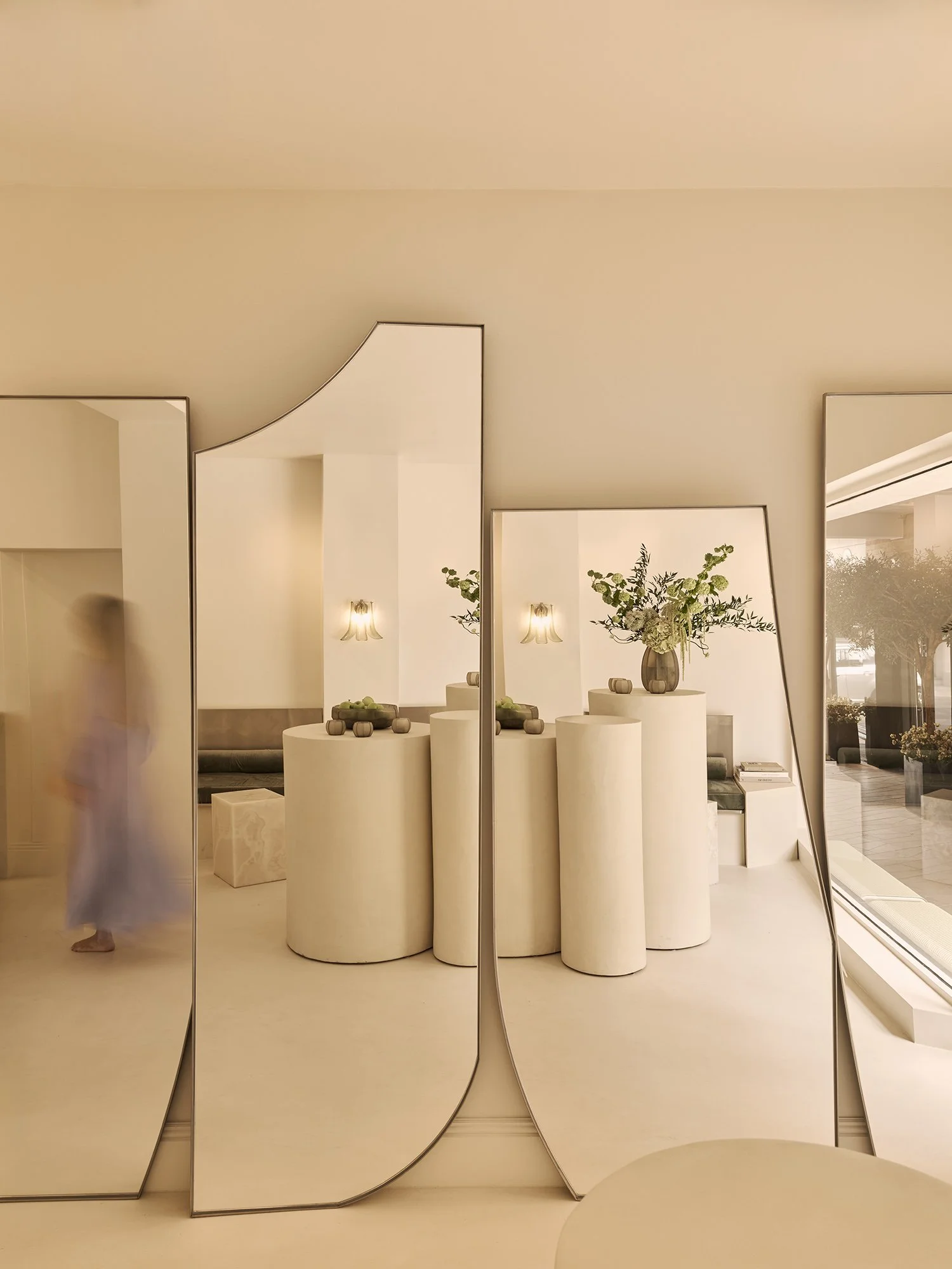 Onyx Matcha Club, London's new destination for matcha and modern rituals. Designed by OT Interiors. Contemporary interior space with curved mirrors, white cylindrical tables, a large vase with green plants, and a beige wall with light fixtures.
