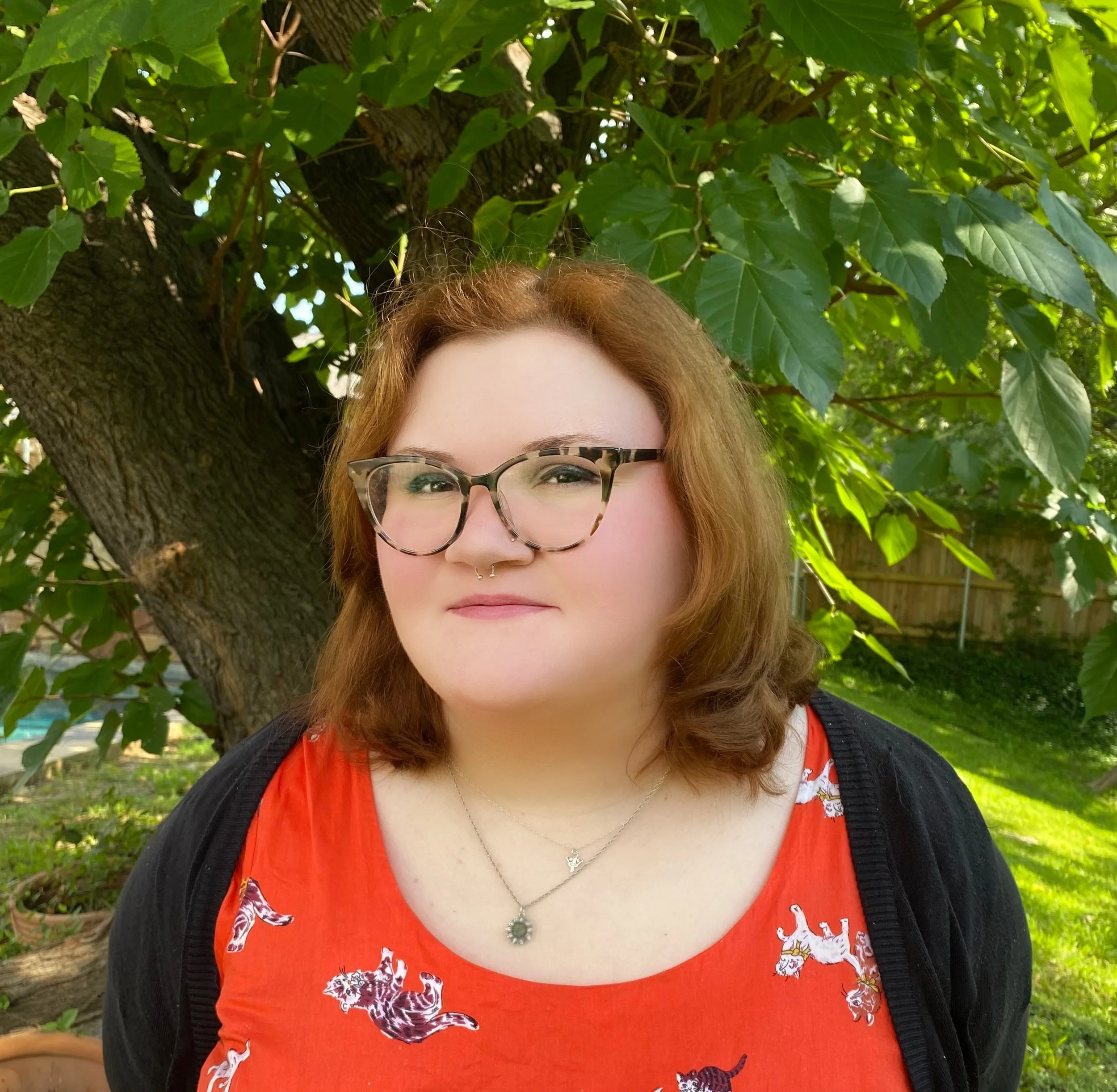 A woman with red hair, glasses, and a septum piercing standing outdoors in front of a large tree with green leaves.