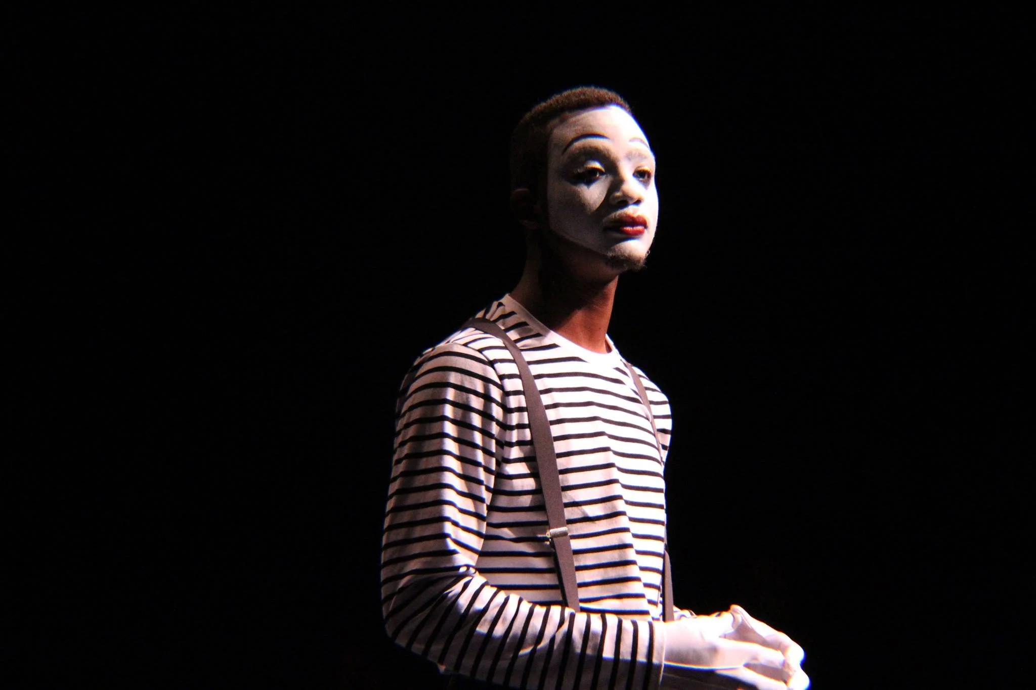 A person with mime makeup, wearing a striped shirt and suspenders, stands on a dark stage with a spotlight on them.