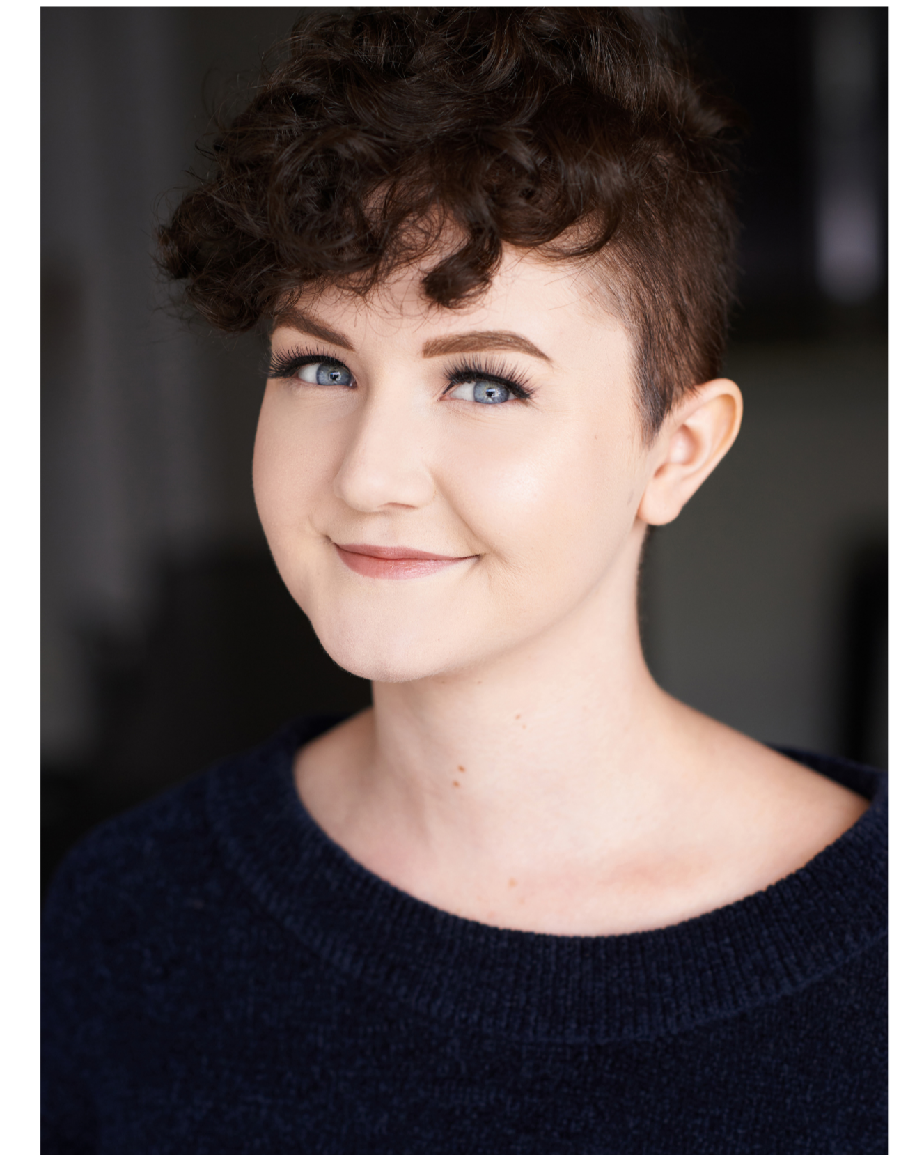 Close-up of a woman with short, curly brown hair, blue eyes, fair skin, and wearing a dark sweater, smiling softly.