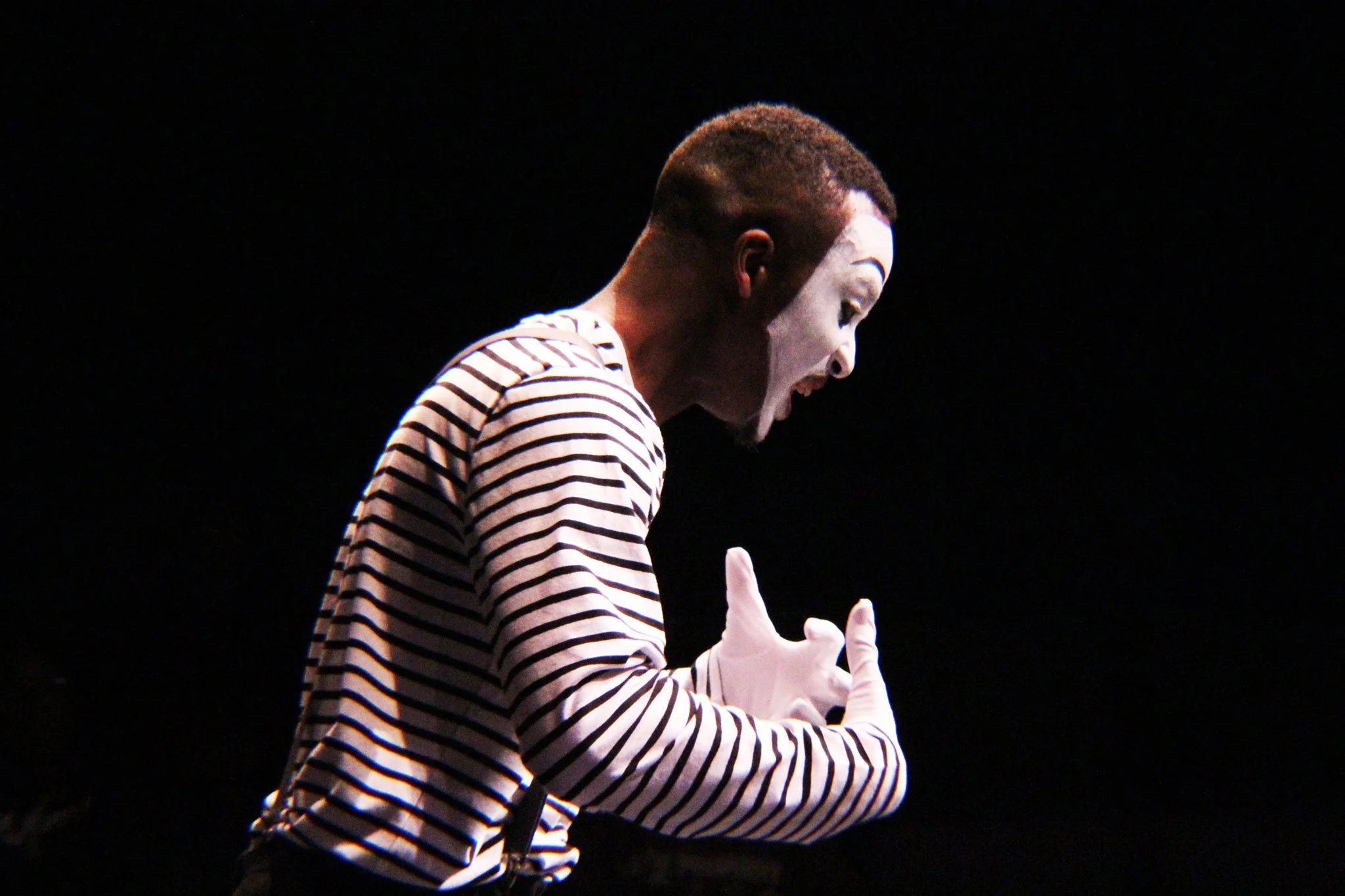 A mime artist dressed in a striped shirt and white gloves, performing on stage with a black background.