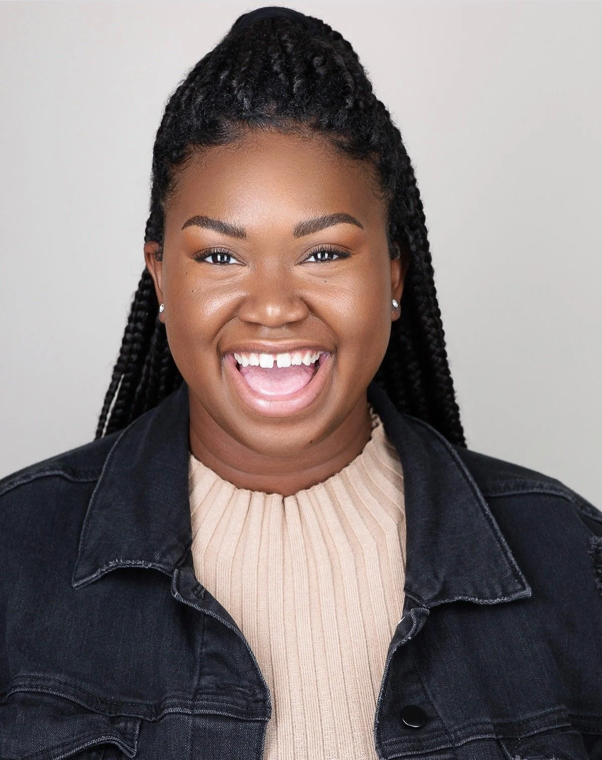 Close-up of a smiling woman with braids, wearing a beige ribbed top and black denim jacket, against a plain light background.