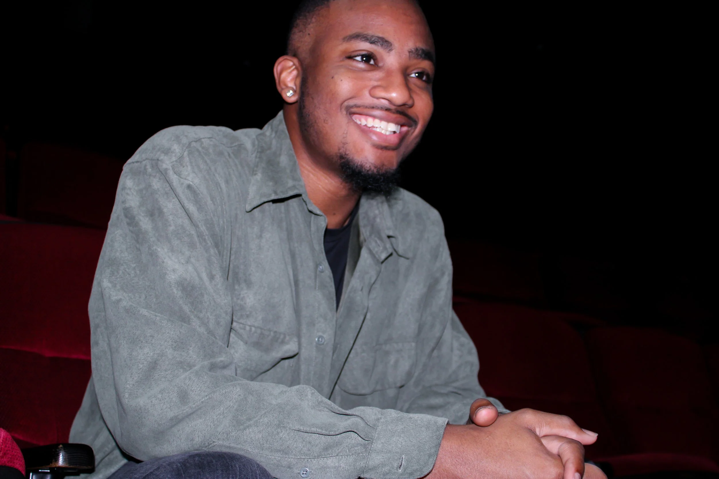 A smiling young man with short hair, earrings, and a goatee, sitting on a dark red seat in a dimly lit environment, wearing a light gray shirt.