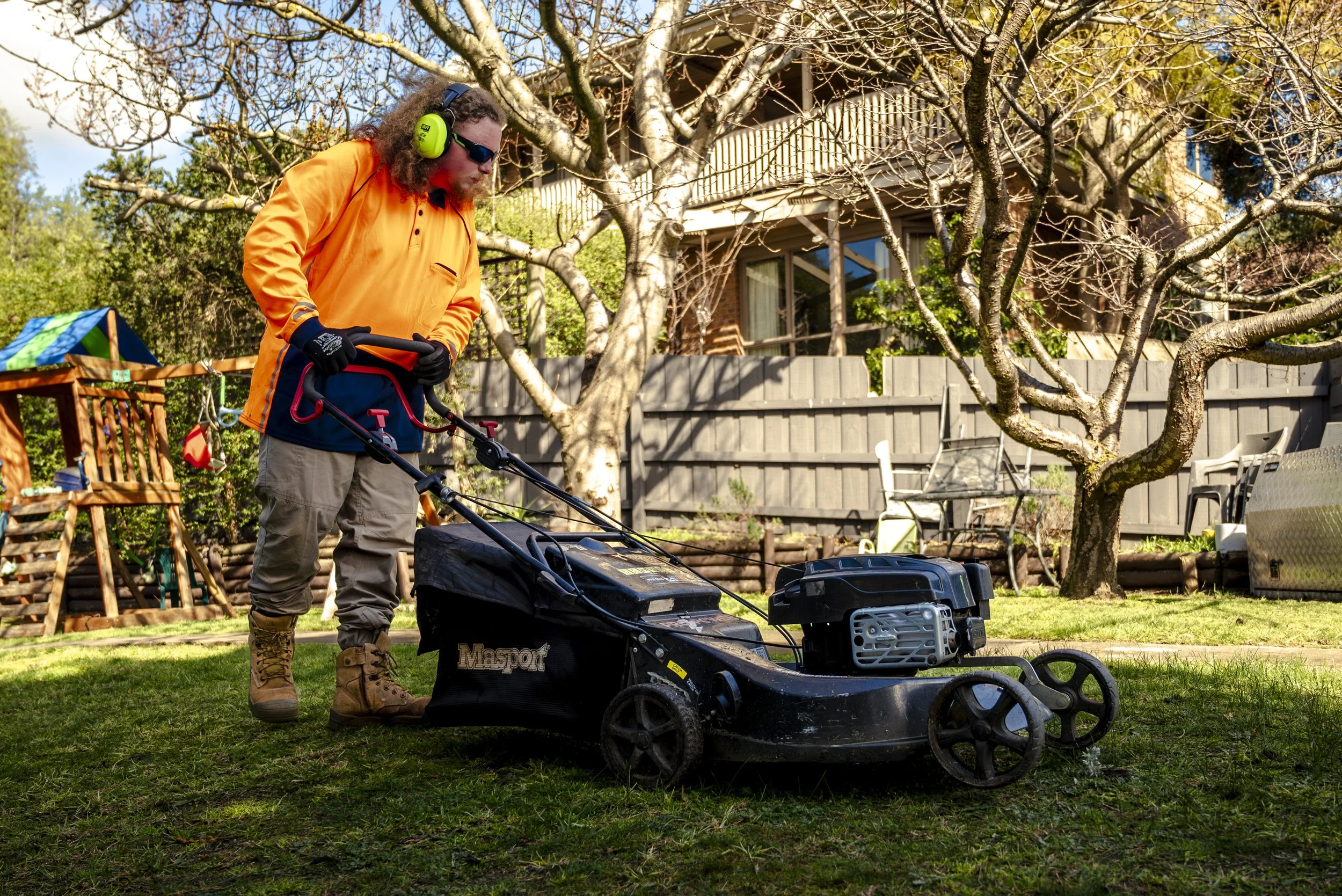 Man in high-vis orange jacket and beige pants mowing lawn with a gas-powered lawn mower in a backyard garden.