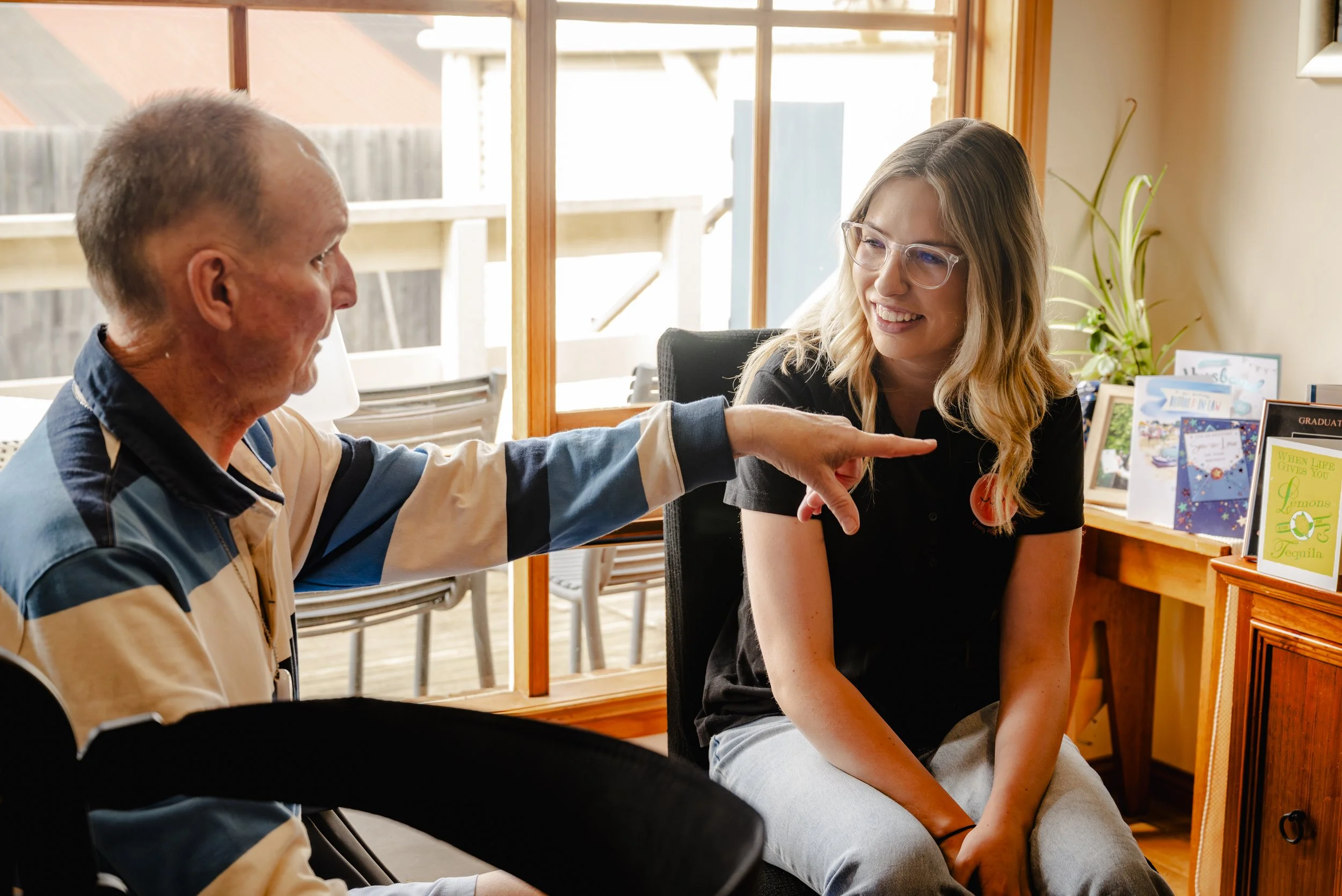 An elderly man pointing at a young woman with Down syndrome sitting at a table, both smiling inside a room with large windows and a wooden cabinet with greeting cards.