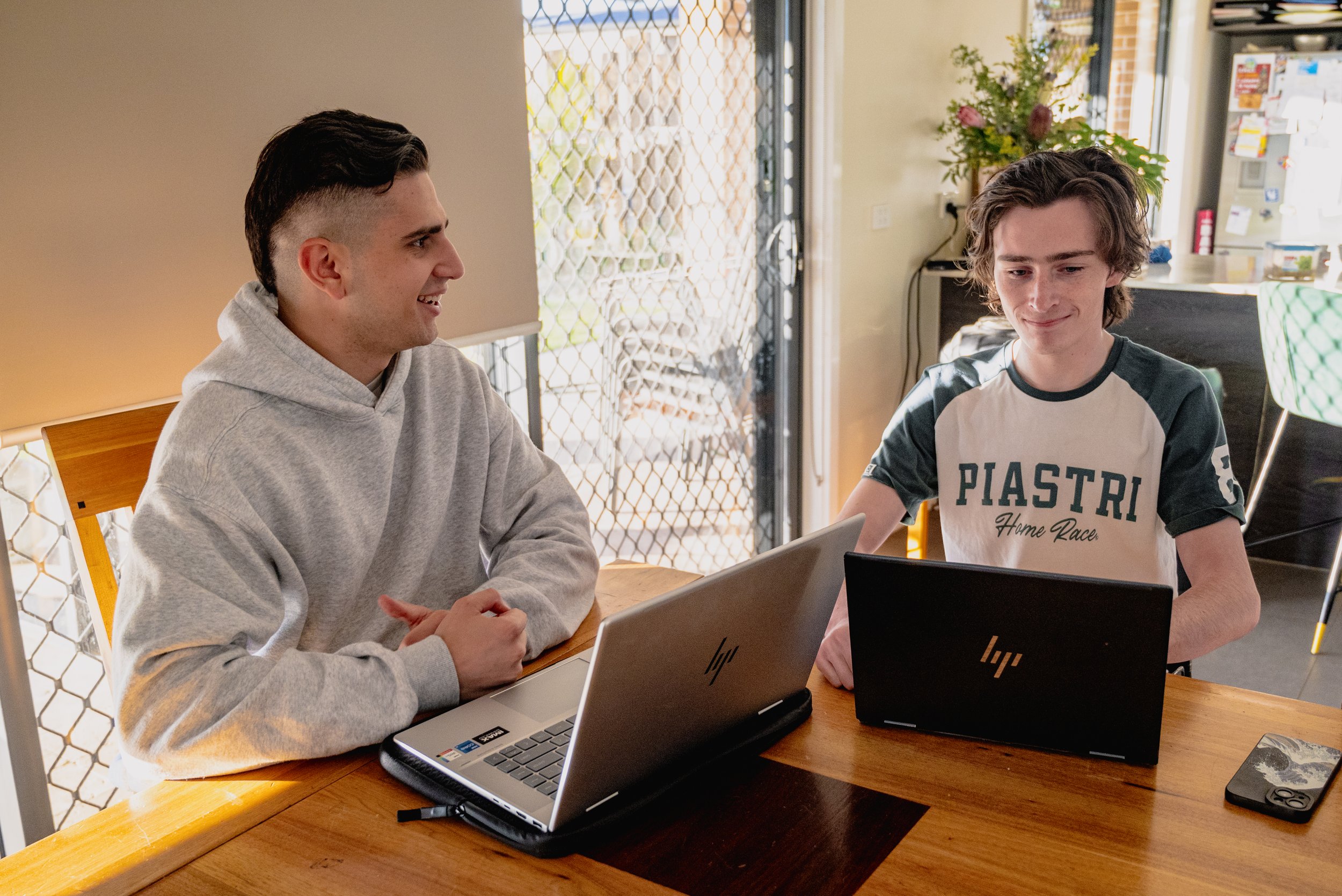 Two young men sitting at a wooden table with laptops, one smiling and wearing a gray hoodie, the other wearing a black and white shirt, with a glass door and plants in the background.