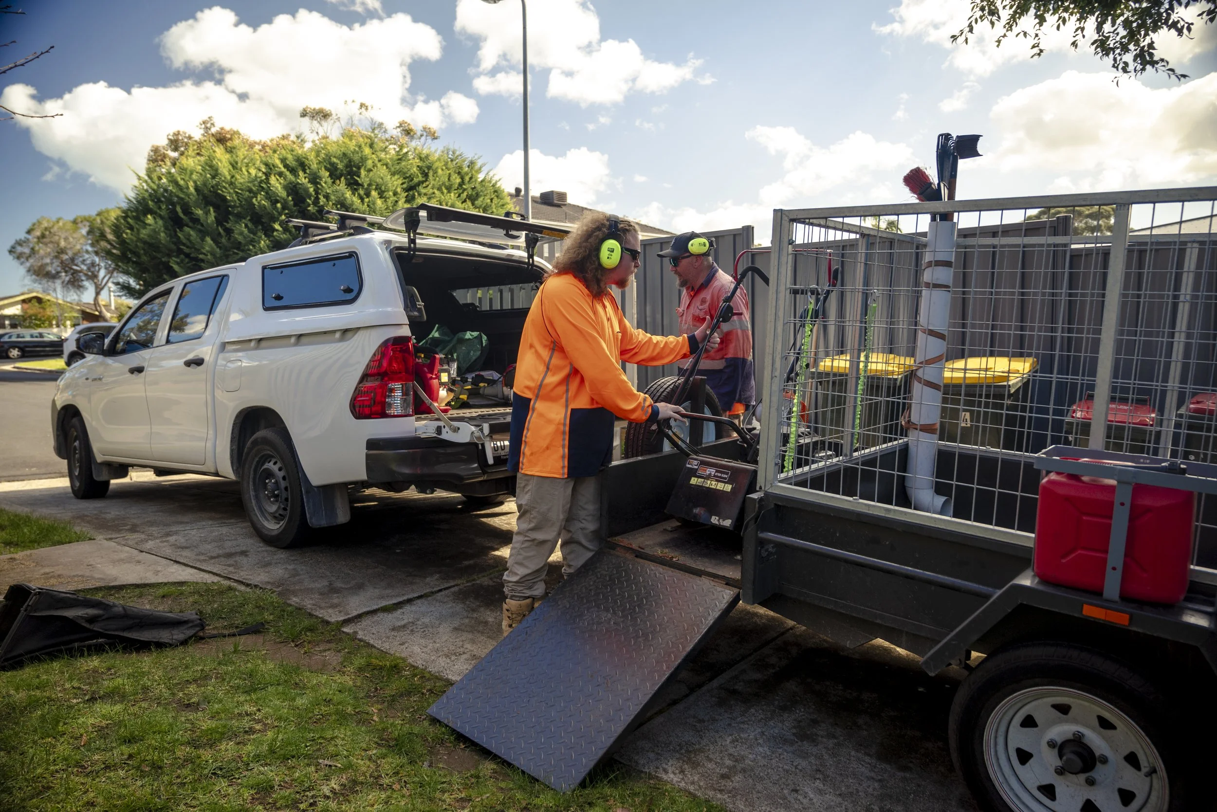 Two workers in orange safety clothing and ear protection are loading equipment into a trailer attached to a white vehicle.