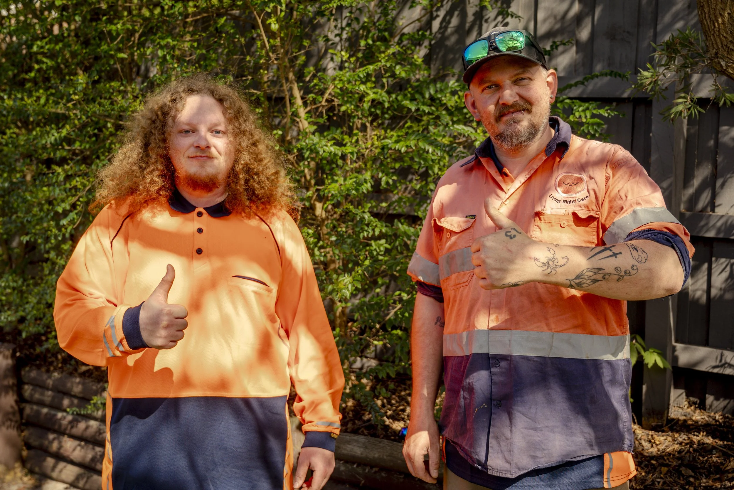 Two men in orange and dark-colored work uniforms standing outdoors in front of greenery and a wooden fence, giving thumbs-up gestures.