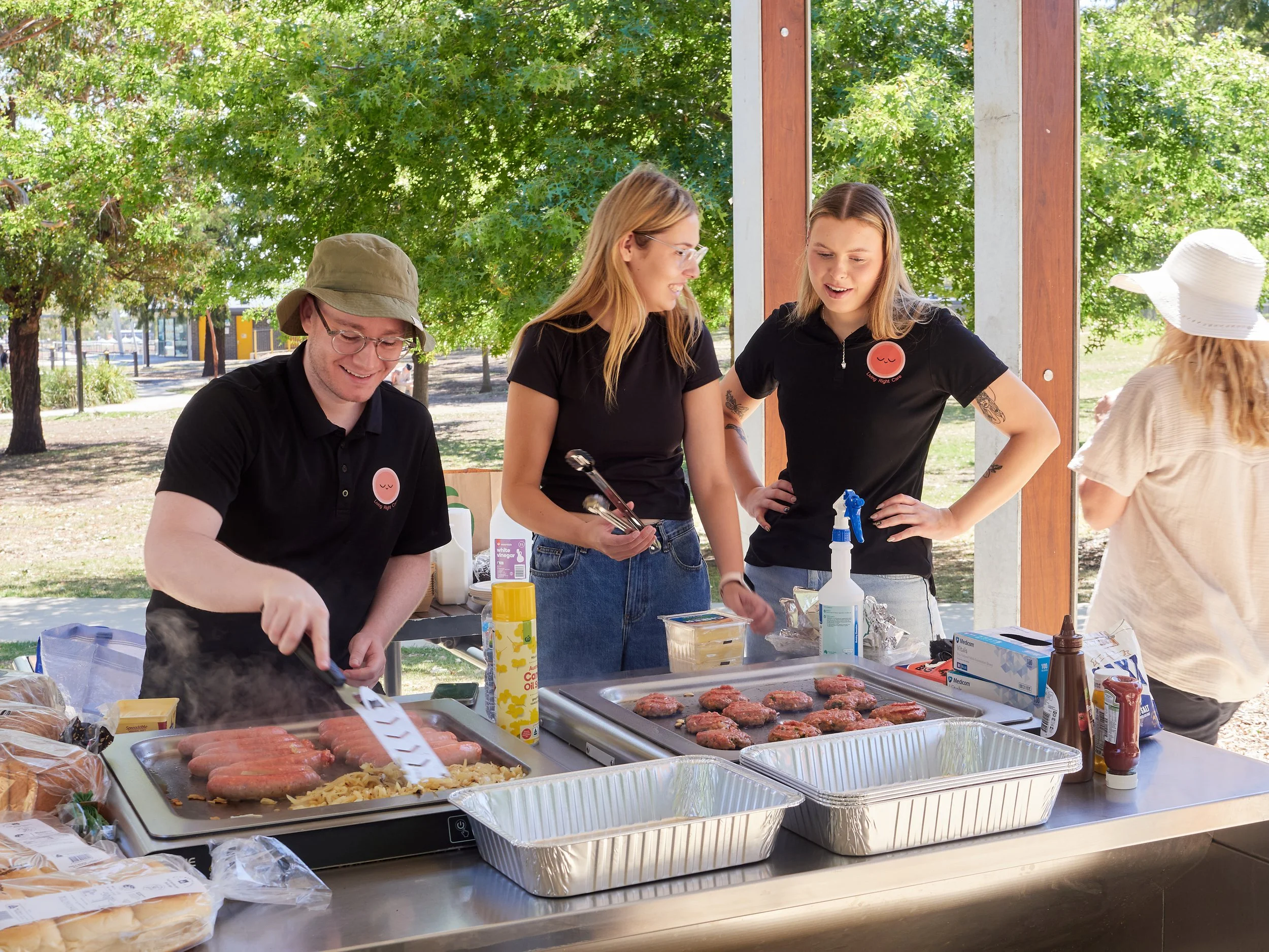 Three young people in Living Right Care branded shirts preparing grilled meat and sausages at an outdoor barbecue station, with trees and a park visible in the background.