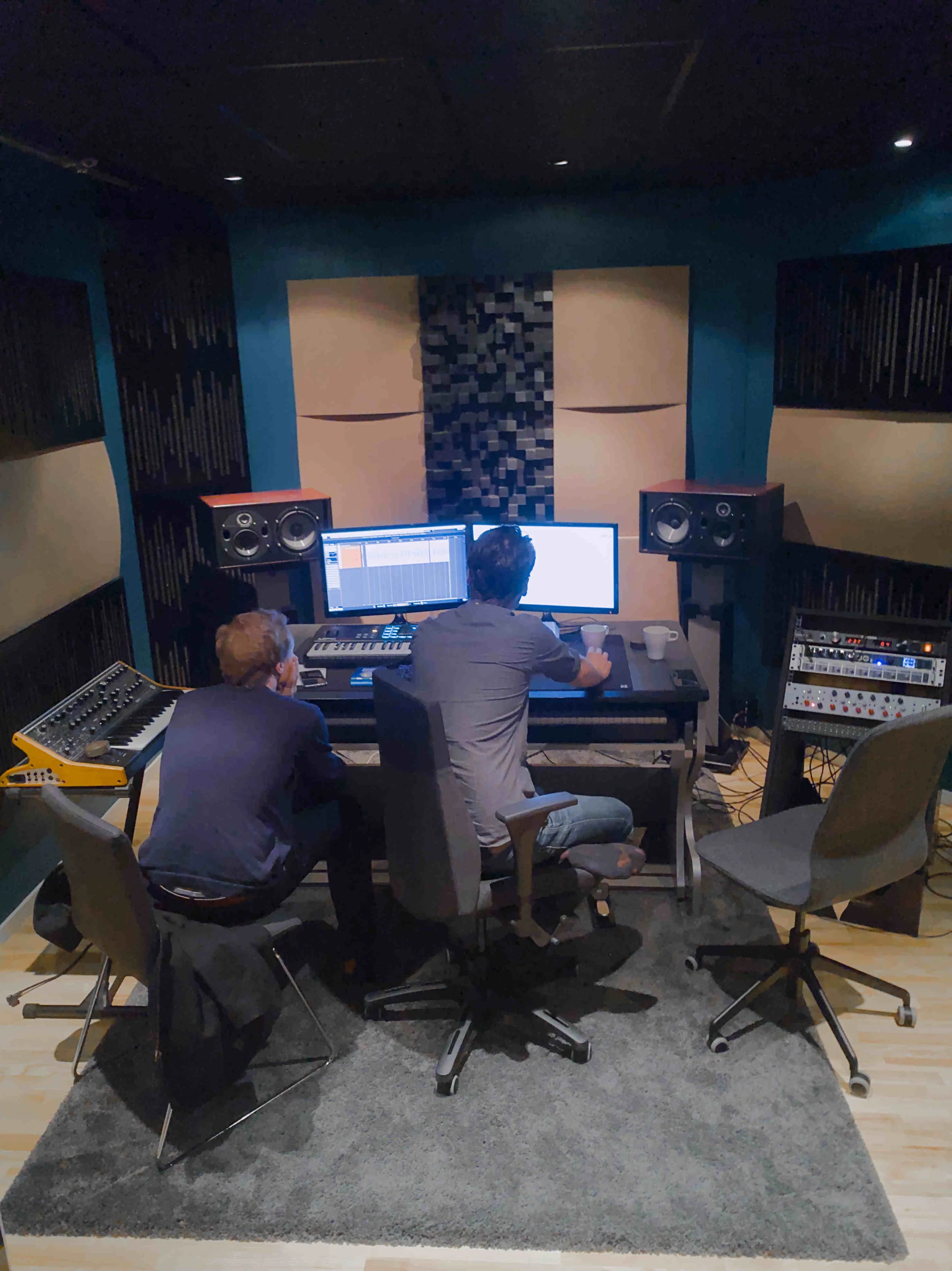 To personer jobber i et innspillingsstudio med to dataskjermer, musikkinstrumenter, lydutstyr og akustiske paneler på veggene.