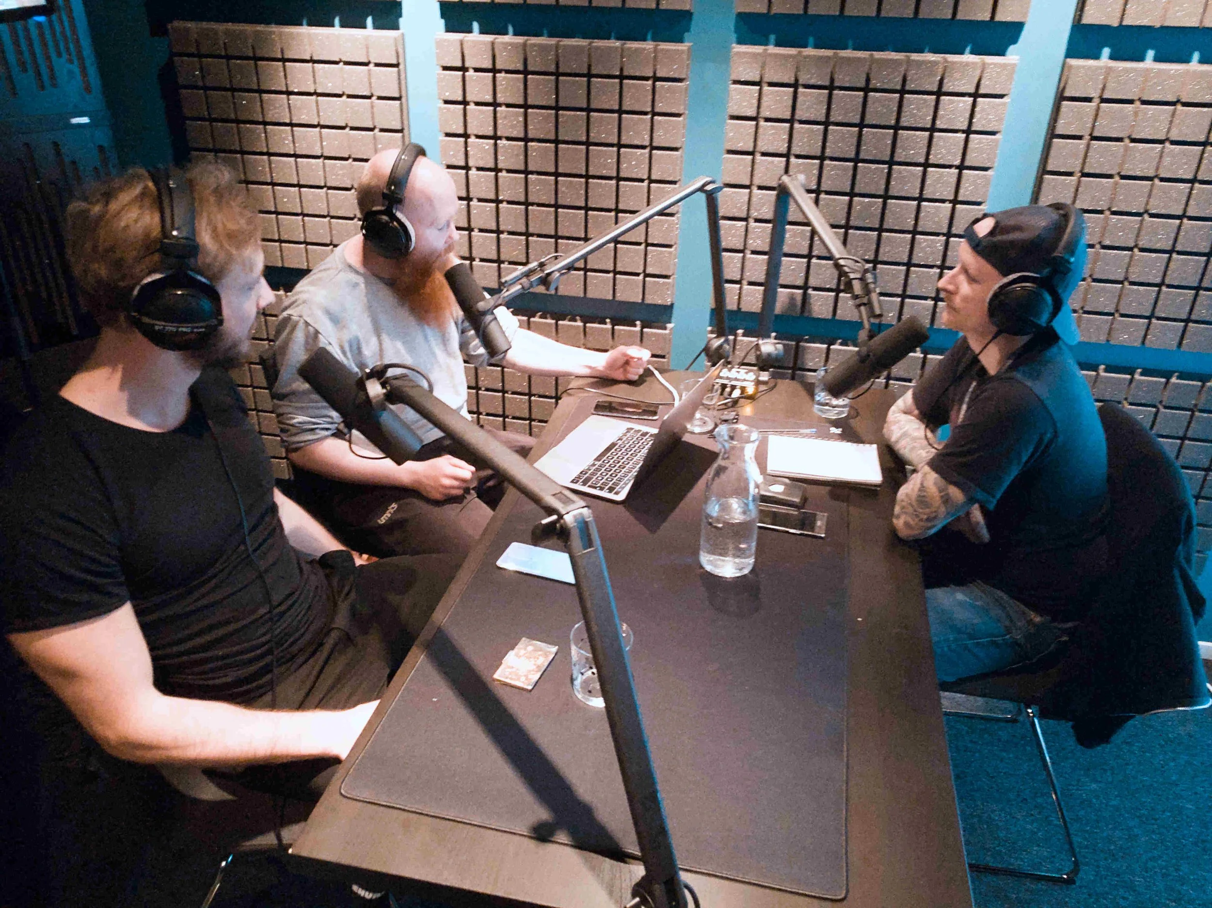 Tre personer i en lydstudio som snakker i mikrofoner. De har hodetelefoner og sitter rundt et bord med laptop og notatblokker.